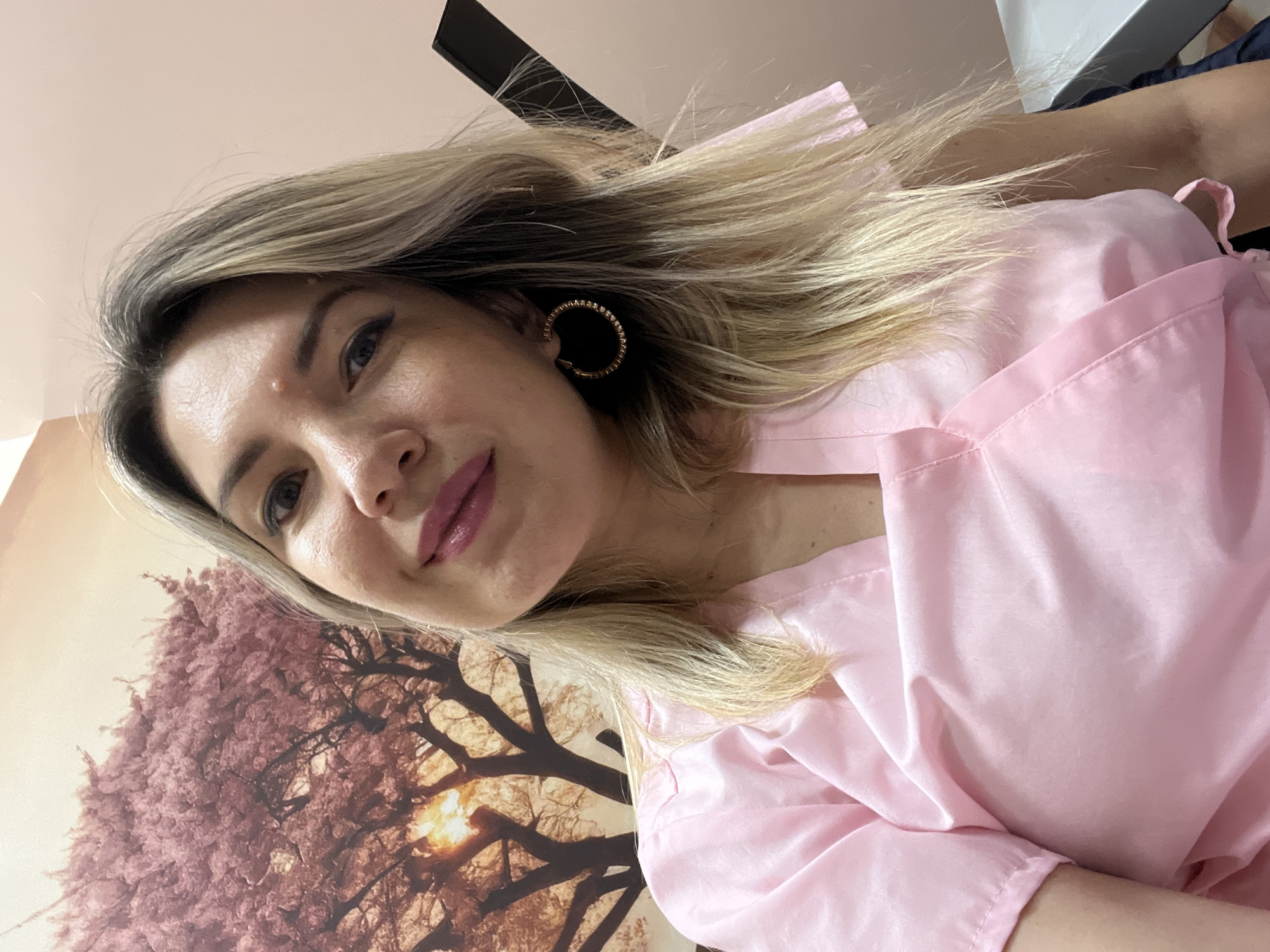 A woman with blonde hair, fair skin, and blue eyes is pictured in a selfie. She is wearing a pink scrub top and large hoop earrings. The background features a wall with a large printed image of a tree with pink foliage. The image is a close-up of the woman's face and upper body. There is no indication of the city of Sochi, Russia.