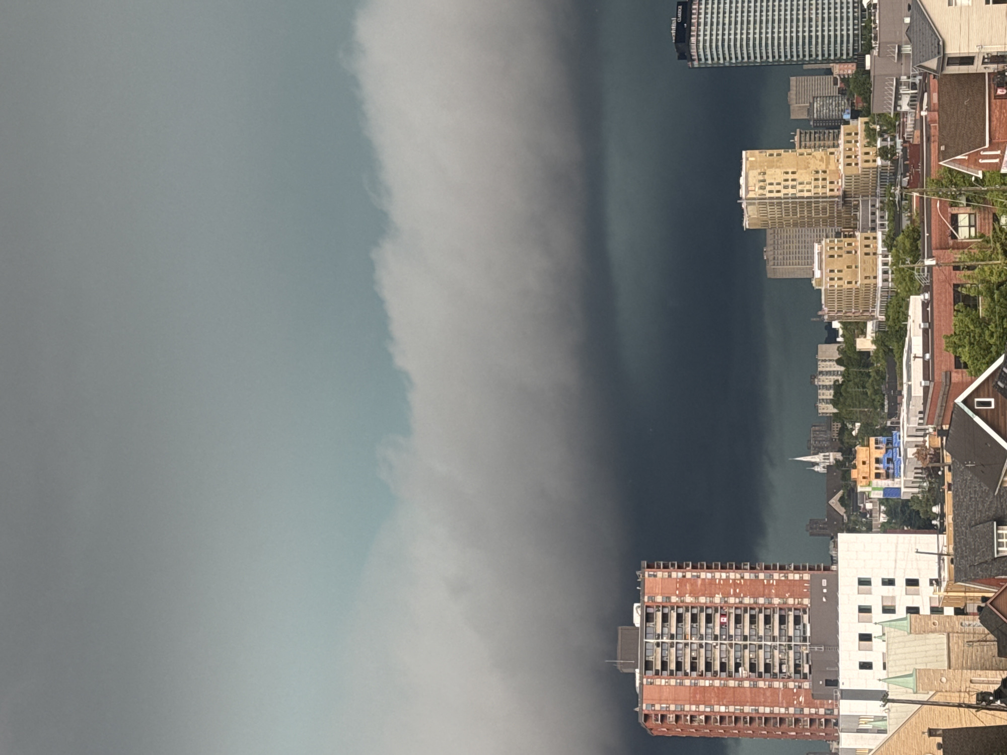 This is a photograph of an urban landscape under a dramatic, dark sky, suggesting an impending storm. The foreground shows a dense collection of buildings, varying in height and architectural style, with rooftops and facades visible. A prominent red-brick, multi-story residential building stands out on the left side of the frame. Several other buildings, including what appear to be churches with steepled roofs, and modern office or apartment complexes, fill the mid-ground and background. The sky is dominated by heavy, dark clouds that create a stark contrast with the lighter, hazy sky above. The overall impression is one of a city preparing for inclement weather. The provided crops confirm details about building structures and cloud formations. The specific location is identified as Ottawa, Canada.