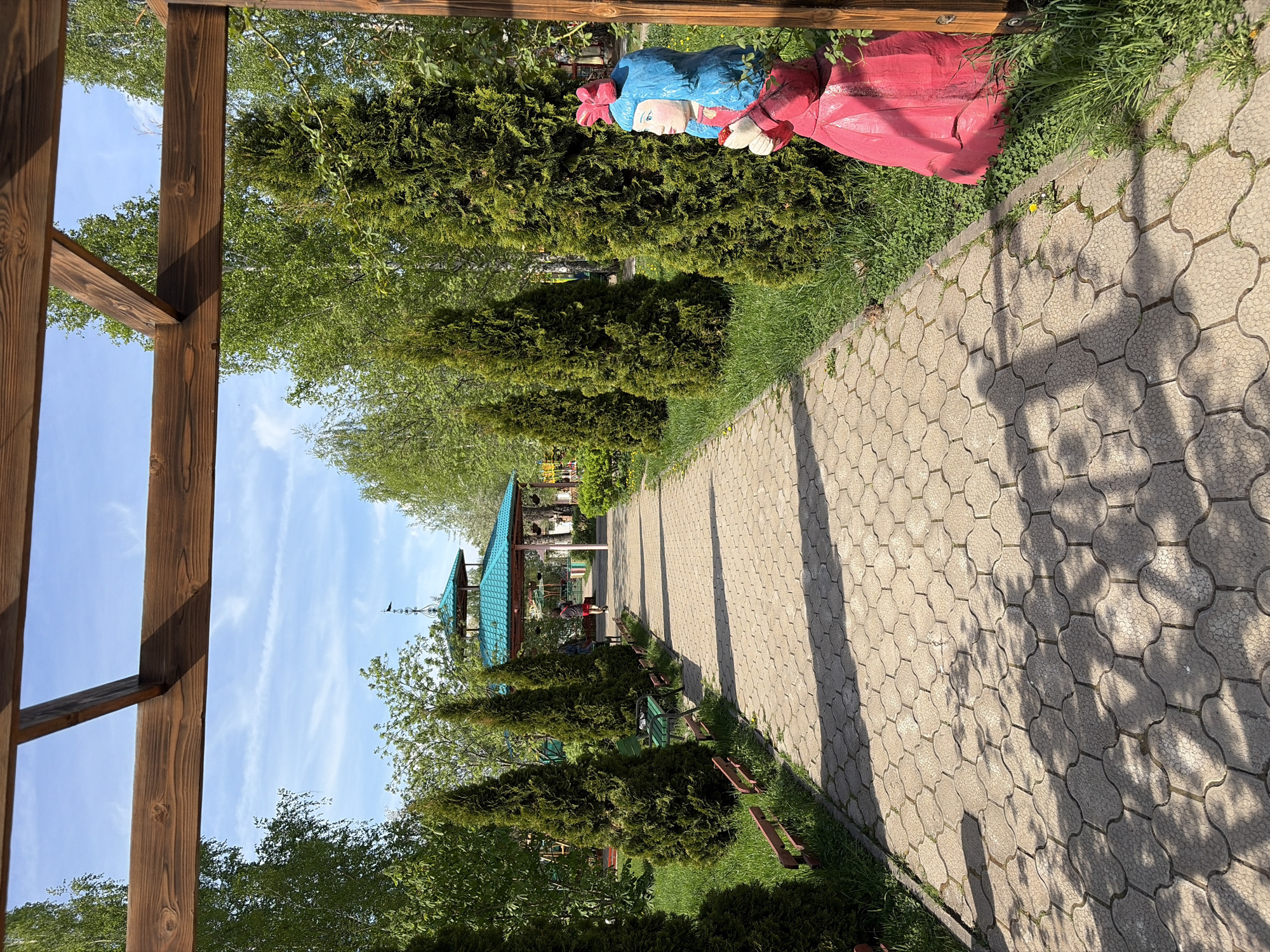The image depicts a paved pathway in a park located in Unknown, Unknown. The pathway is made of interlocking grey paving stones, with shadows from trees casting patterns on its surface. Along the pathway are neatly trimmed evergreen shrubs and patches of grass. To the right of the path, a statue of a woman in a red dress and blue head covering stands amidst the greenery. In the background, a gazebo with a blue tiled roof is visible, with a few people underneath it. The sky is blue with wispy clouds. In the foreground, a wooden structure, possibly part of a pergola or gate, frames the left side of the image.