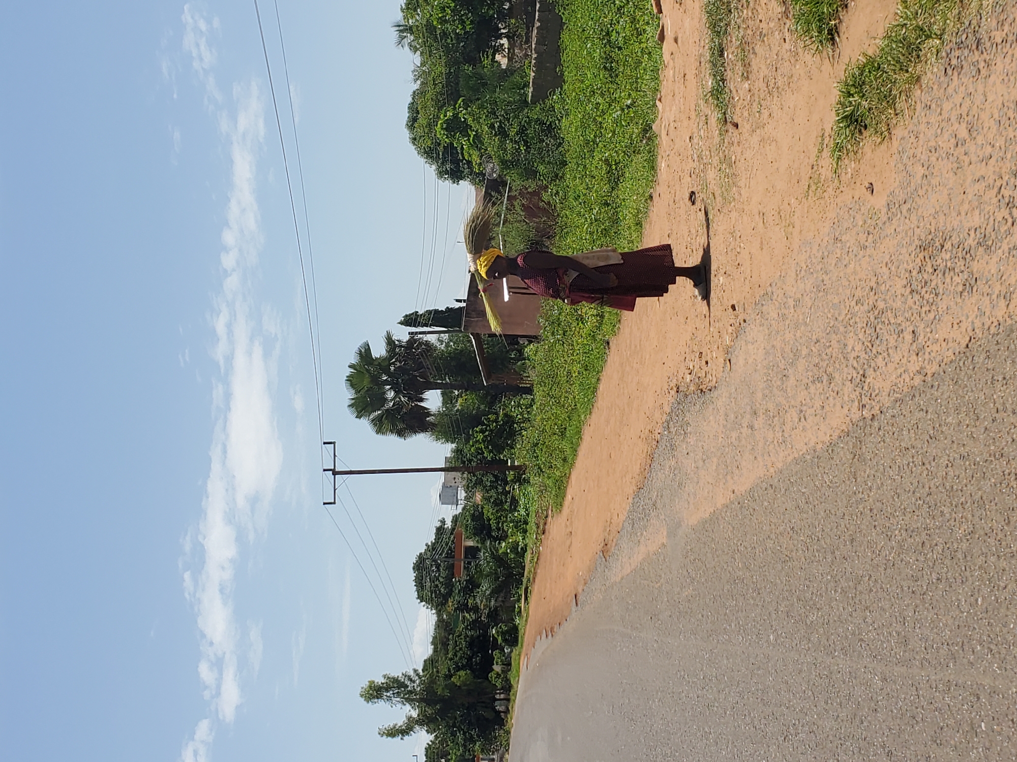 A woman wearing a yellow headwrap and a red patterned dress stands on the side of a dirt road, balancing a bundle of straw on her head. She is seen in profile, looking to the left. The road is next to a grassy area with a cluster of trees and buildings in the background. A utility pole with power lines is visible on the right side of the frame. The sky above is clear blue with a few scattered white clouds.