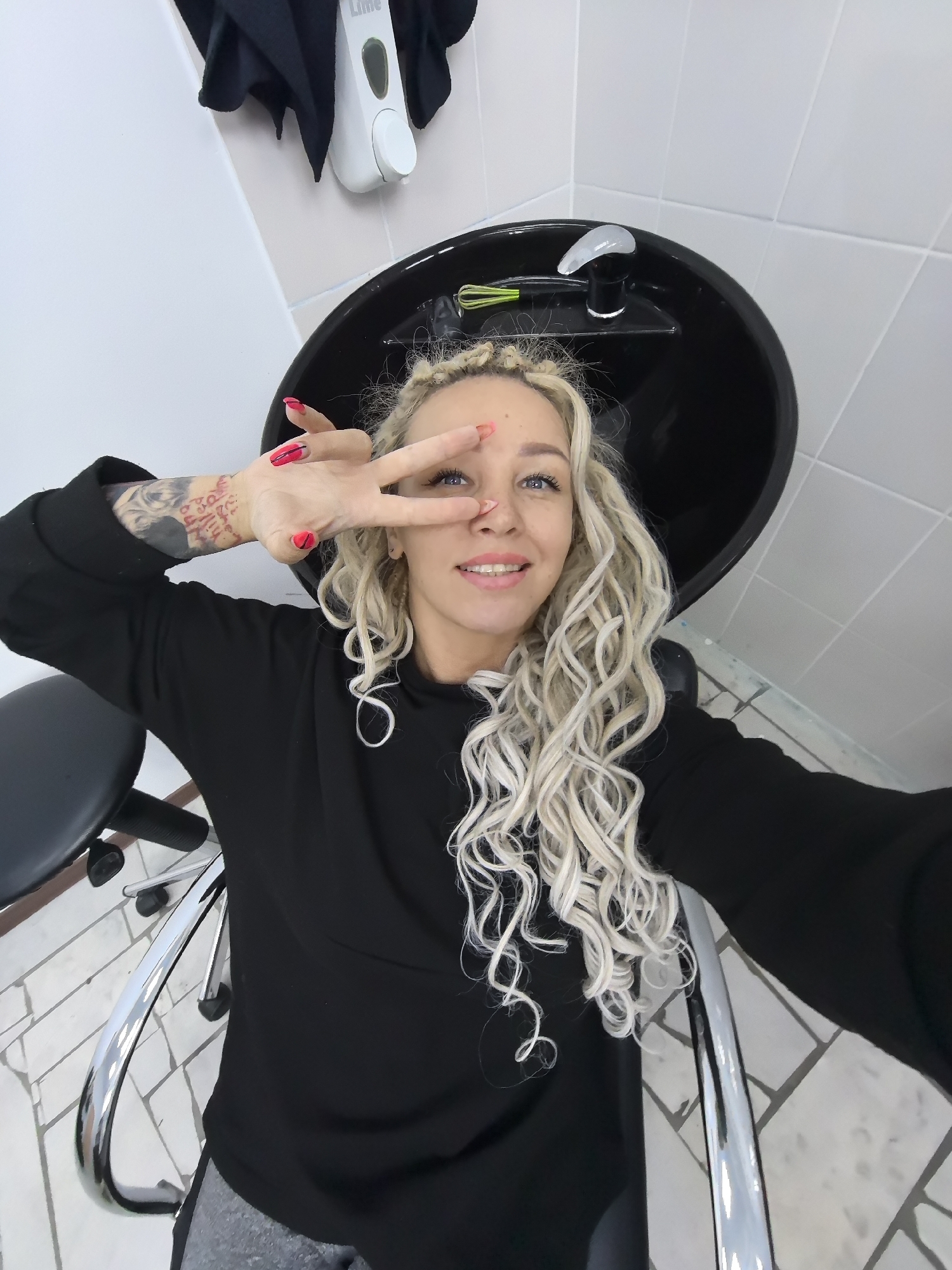 In the city of Chaikovsky, Russia, a young Caucasian woman with blonde curly hair and braids, wearing a black sweatshirt and gray pants, sits in a black chair in what appears to be a hair salon. She is making a peace sign with her right hand, with bright red nail polish visible. She has a tattoo on her left arm and is looking at the camera with a smile. Behind her, a black sink with a faucet is visible, and the walls are tiled in white. A black towel hangs on the wall to the left of the sink. The floor is tiled with a pattern of light and dark gray squares.