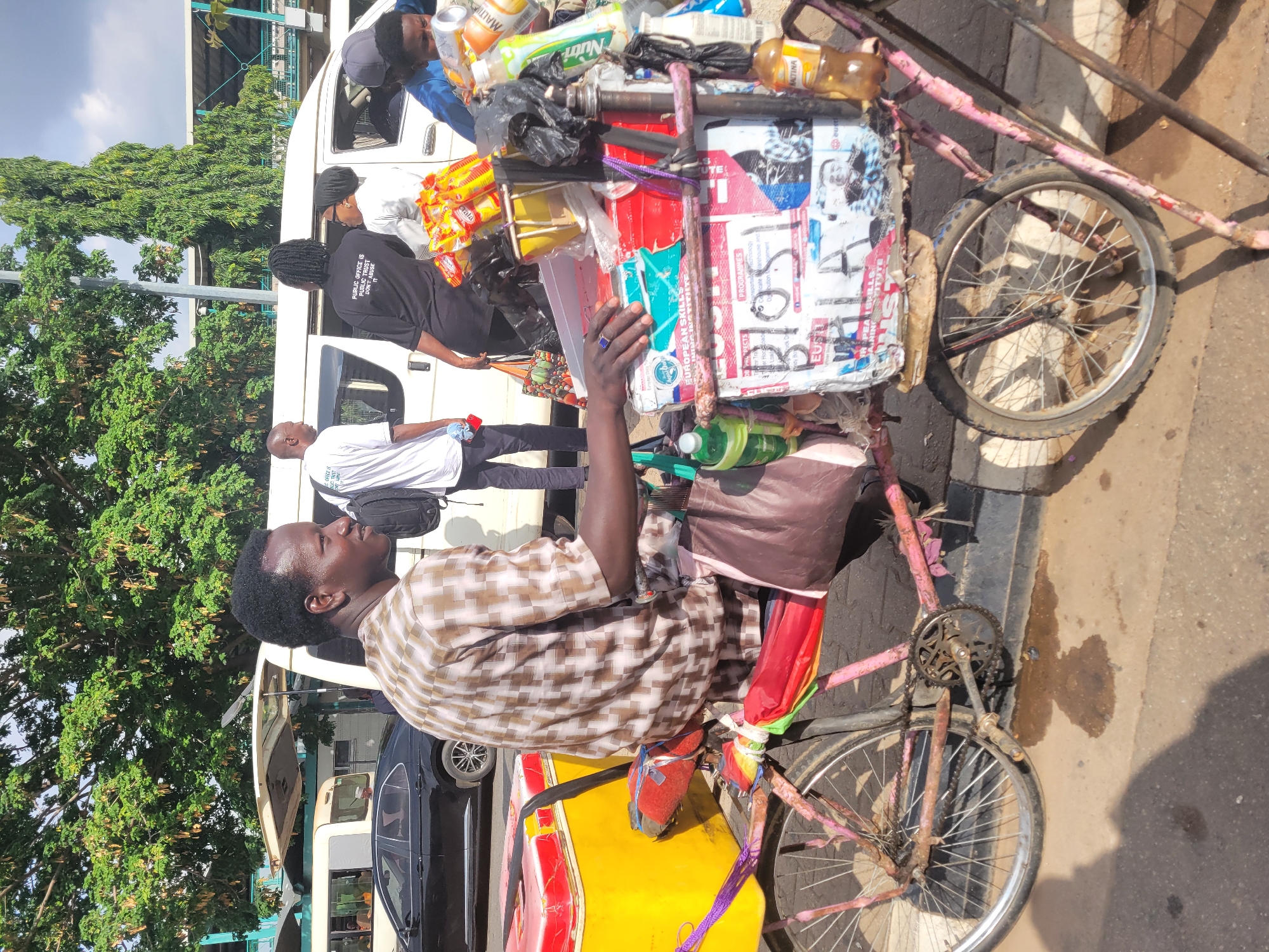 This image depicts a vibrant street scene in Abuja, Nigeria, during daylight hours under sunny weather. The central subject is a man, likely a street vendor, seated on a modified tricycle. He has dark skin, short dark hair, and wears a brown and white checkered top along with a patterned brown wrapper or skirt. He holds a clear plastic cup in his hand, looking slightly towards the right.
The tricycle itself features a faded pink/red frame and a prominent yellow and red cargo box at the front. This box is heavily loaded with various goods for sale, including plastic bottles of beverages such as "Maltina" and "Nutri-Milk," packaged snacks, and other items contained within a black plastic bag and a yellow drum-like object. The side of the cargo box is adorned with torn posters and advertisements, displaying fragmented text like "BURO," "EUS," "PROGRAMMES," "PEA SKILLS," and "EUROPEAN INSTITUTION."
In the background, a white passenger van, possibly a commercial minibus, is parked. Several individuals are seen near the van; one person wears a black t-shirt with the visible text "PUBLIC OFFICE IS PUBLIC TRUST DON'T ABUSE IT," while another is in a white t-shirt. Lush green trees line the street, and a portion of a black car is visible. The asphalt ground shows distinct shadows, confirming the bright, sunny conditions. The scene captures a moment of daily commerce and urban life.