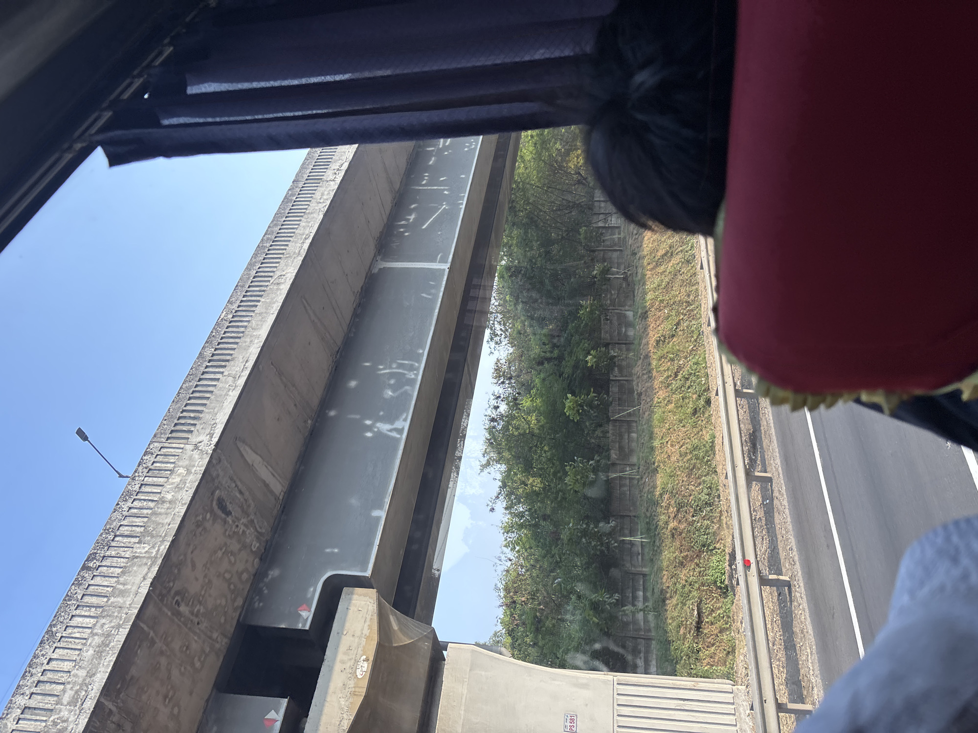 The image shows a view from inside a vehicle looking out at an elevated highway structure. The highway is made of concrete and steel, with a gray railing along the side. Below the highway, there are trees and a concrete wall. A person's head is visible in the foreground, and a red seat is in the bottom right corner. A single red object, possibly a flower, is on the railing. The sky is blue. The photo was taken in Karawang, Indonesia.