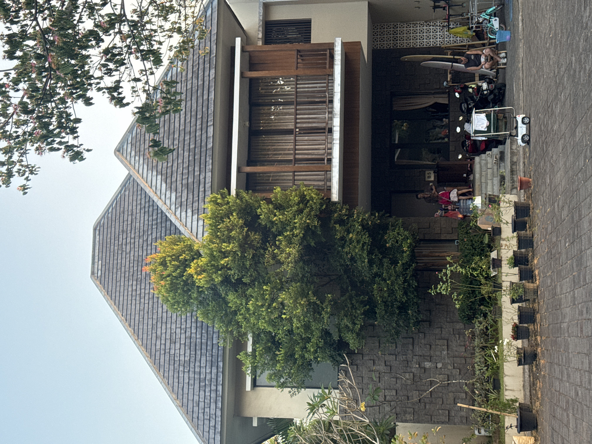 This is a full-shot exterior, eye-level photograph of a two-story house with a multi-faceted, dark gray tiled roof, located in Kuta Selatan, Indonesia. The house features a prominent stone facade on the lower level and a wooden-accented balcony on the upper level. A large green tree with vibrant leaves is positioned in front of the house, partially obscuring the lower windows. Several potted plants are arranged on the paved driveway and in front of a low garden wall.
On the driveway, two motorcycles are parked. To the right of the motorcycles, a man is seated on a small white stool, looking down at something in his hands. Next to him, a turquoise bicycle is leaning against a rack. A blue bucket sits on the ground near the bicycle.
In front of the house entrance, a woman stands on the steps holding a pink bag. To her left, a young girl in a white skirt and pink top is seated, and a black cart with white clothes draped over it is positioned in front of her. A small white toy car is on the driveway in front of the cart.
The environment suggests a warm climate with bright sunlight casting shadows. The scene appears casual and relaxed, with everyday objects and people present.
