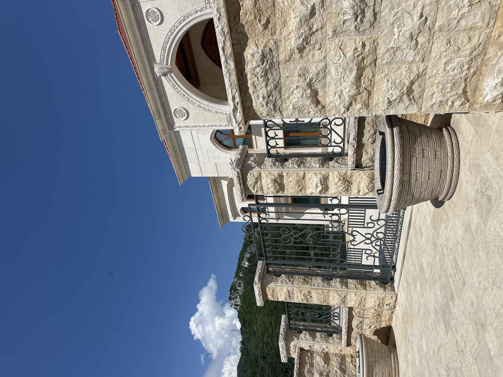 An outdoor, eye-level, wide shot captures a stone facade, ornate metal gate, and lush green mountain scenery under a bright blue sky with scattered clouds. The building's exterior, constructed from light-colored stone blocks, features decorative arches and windows with wooden frames. A black wrought iron gate, adorned with intricate scrollwork, stands partially open, leading to an unseen courtyard. Flanking the gate are two large, textured planters. In the background, a dense forest covers a steep mountainside, and a single, fluffy white cloud drifts across the sky. The foreground is paved with light-colored stone tiles. The scene is devoid of any people or animals. This appears to be a view of the Asen's Fortress in Asenovgrad, Bulgaria, a historical landmark perched on a rocky outcrop overlooking the Rhodope Mountains.