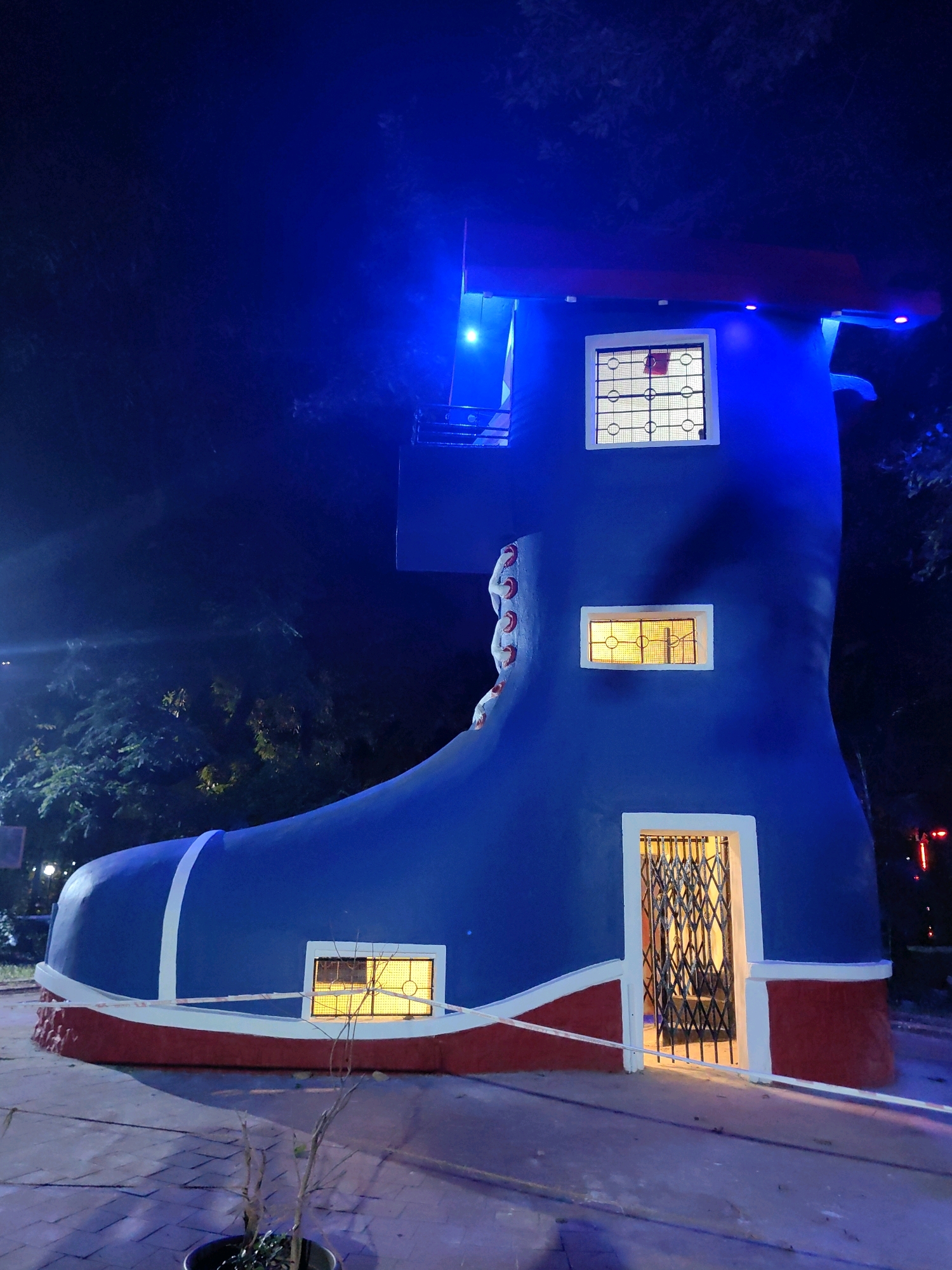 The image shows a large, blue boot-shaped structure at night. The structure has red trim along the bottom and white trim along the top of the sole. There are three windows visible, each with decorative grilles and illuminated from within. The lowest window is near the entrance, which is a doorway with a metal security gate. The structure is lit with blue lights, casting a blue glow on the surrounding dark environment. Trees are visible in the background. The ground in front of the structure appears to be paved. The structure is located in the city of Mumbai, India, and is part of the "Giant Boot" attraction at the Byculla Zoo.