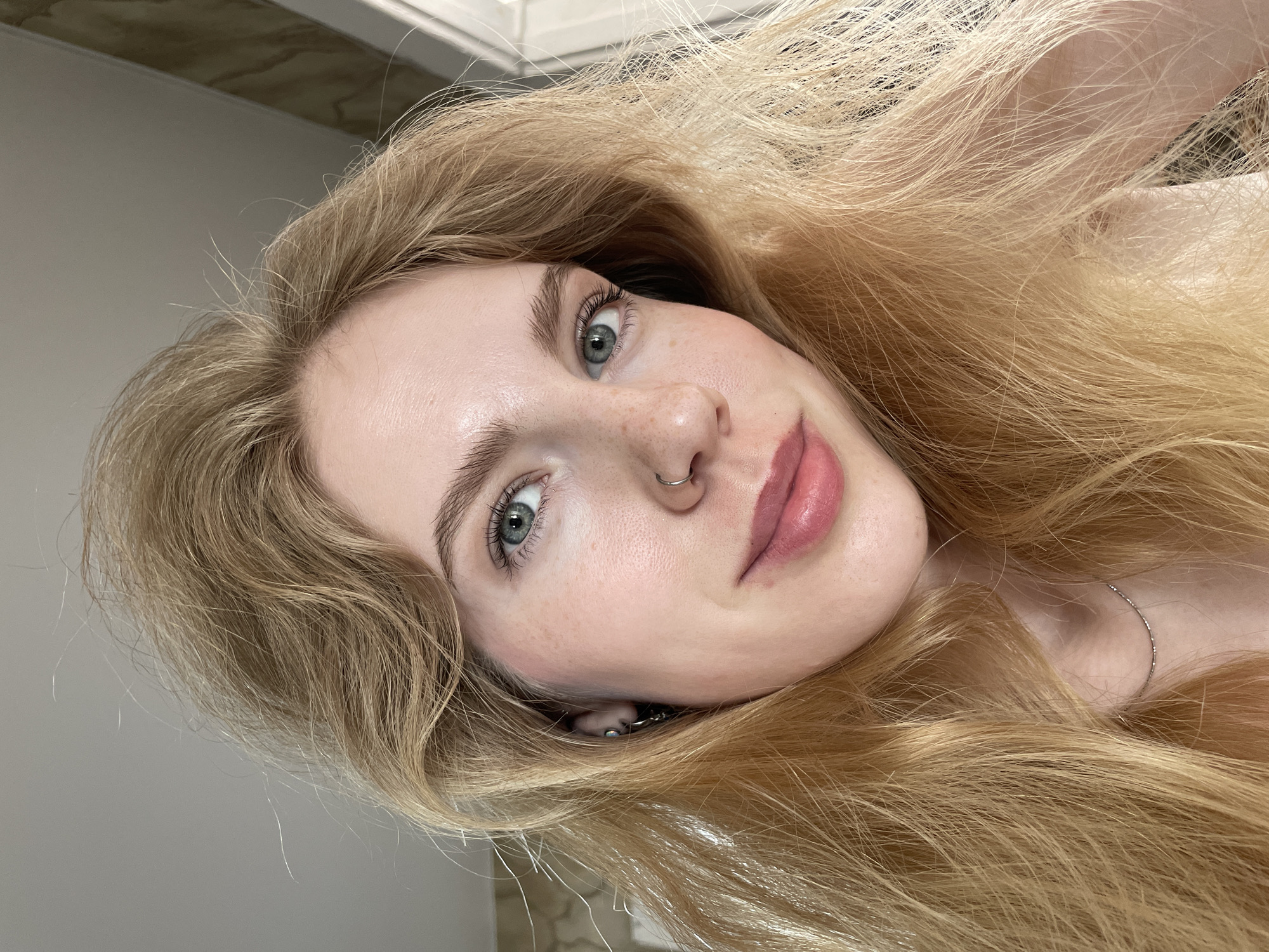 A close-up, tilted portrait captures a fair-skinned person with long, blonde hair that is flowing around their face. The person has light blue eyes, freckles on their nose and cheeks, and a piercing in their left nostril. They are wearing a delicate silver necklace. The background consists of a white or light-colored wall and a partially visible ceiling corner. There is no information available in the image regarding the city of Izhevsk, Russia.