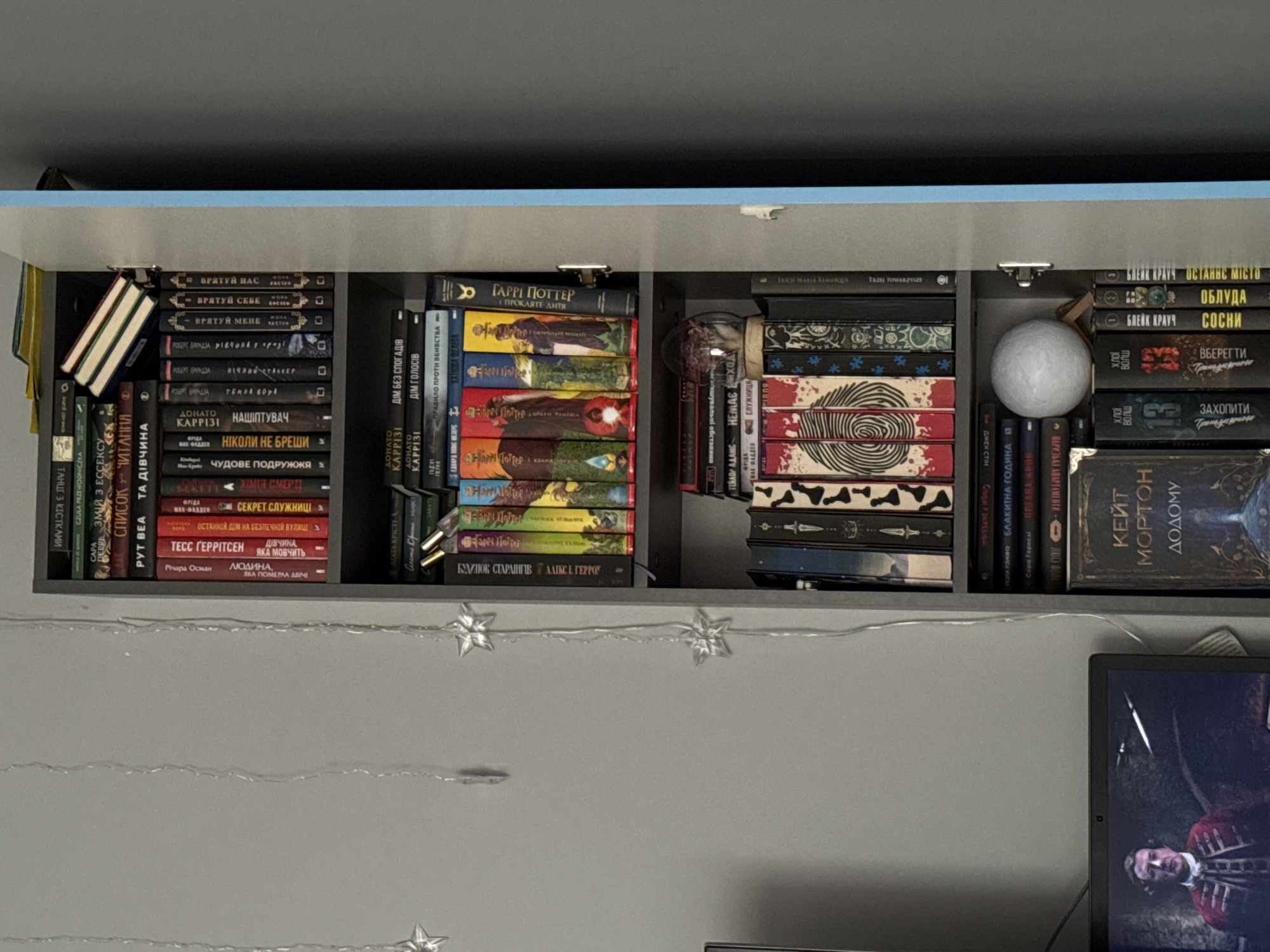 The image depicts an open, dark gray tall cabinet filled with books. The cabinet is positioned against a plain, light gray wall. Several strings of fairy lights with star-shaped ornaments hang in front of the cabinet on the left side. The cabinet has several shelves, each containing books arranged vertically. Some books are visible from their spines, displaying titles and authors in various languages, predominantly Cyrillic script.
On the top shelf, a small stack of colorful books is leaning against the back of the cabinet. The second shelf from the top holds a row of books, some with prominent gold lettering. The third shelf displays more books, including some with illustrations on their spines. The fourth shelf contains a collection of books with varied spine designs, and a small glass sphere is placed on top of some of them. The bottom shelf is packed with more books, and a large, dark-covered book with gold lettering is prominently displayed.
To the left of the cabinet, a laptop screen is partially visible, showing an image of a person in historical attire. The overall scene suggests a domestic or personal space, likely a room where books are stored and displayed. There is no identifiable information in the image that would suggest a location within the city of Łódź, Poland, nor are there any people actively engaged in actions or interactions. The environment is static and focused on the display of books.