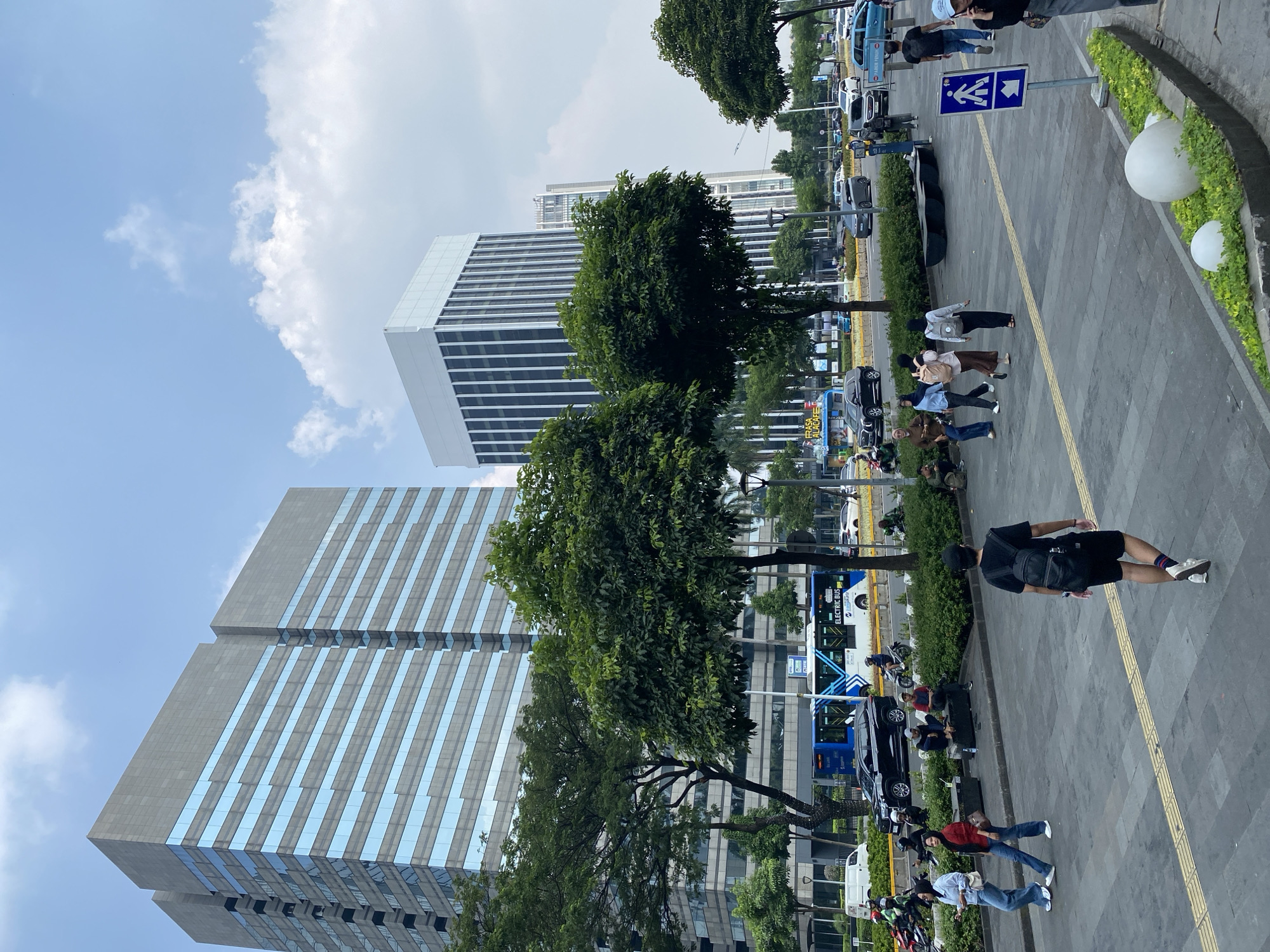 The image depicts a street scene in Central Jakarta, Indonesia. Two modern, tall buildings dominate the background, one with a gray facade and blue glass windows, the other with a white facade and vertical black window frames. In the middle ground, an electric bus with blue and white graphics is visible, alongside other vehicles including cars and motorcycles, on a busy street. Lush green trees line the street and a pedestrian walkway. Several people are seen walking on the walkway, some are seated on benches. One person is walking away from the camera, carrying a black backpack. Another group of people, including women in headscarves and jeans, are walking towards the left side of the image. In the foreground, a yellow textured tactile paving indicates a pedestrian crossing. Decorative white spheres and green plants are arranged along the edge of the walkway. The sky is partly cloudy with a bright blue hue.