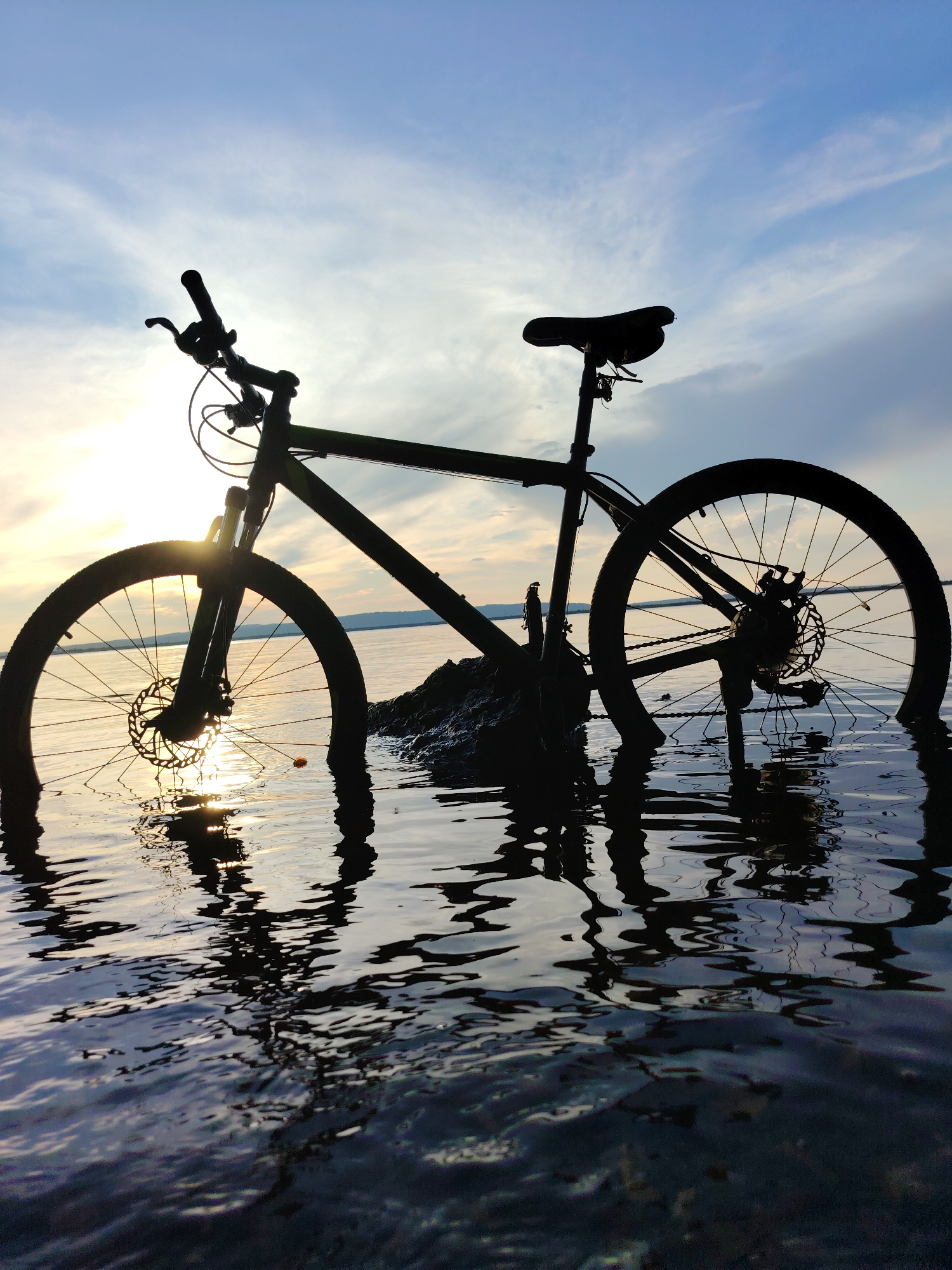 An outdoor, low-angle, full shot captures a bicycle silhouette submerged in shallow water, with its tires and bottom half of the frame partially immersed. The bicycle is dark and positioned near a rocky area. The sky above is a gradient of blue and white, with streaks of clouds, and the sun is visible as a bright, hazy orb peeking through from the left, casting reflections on the water's surface. The scene appears to be a body of water, possibly a river or lake, with a faint horizon line of hills in the distance. The water is calm, with gentle ripples reflecting the sky and the submerged bicycle. The image conveys a sense of quiet and solitude. No people are present in the scene. The location is the Kama River in Chaikovsky, Russia.
