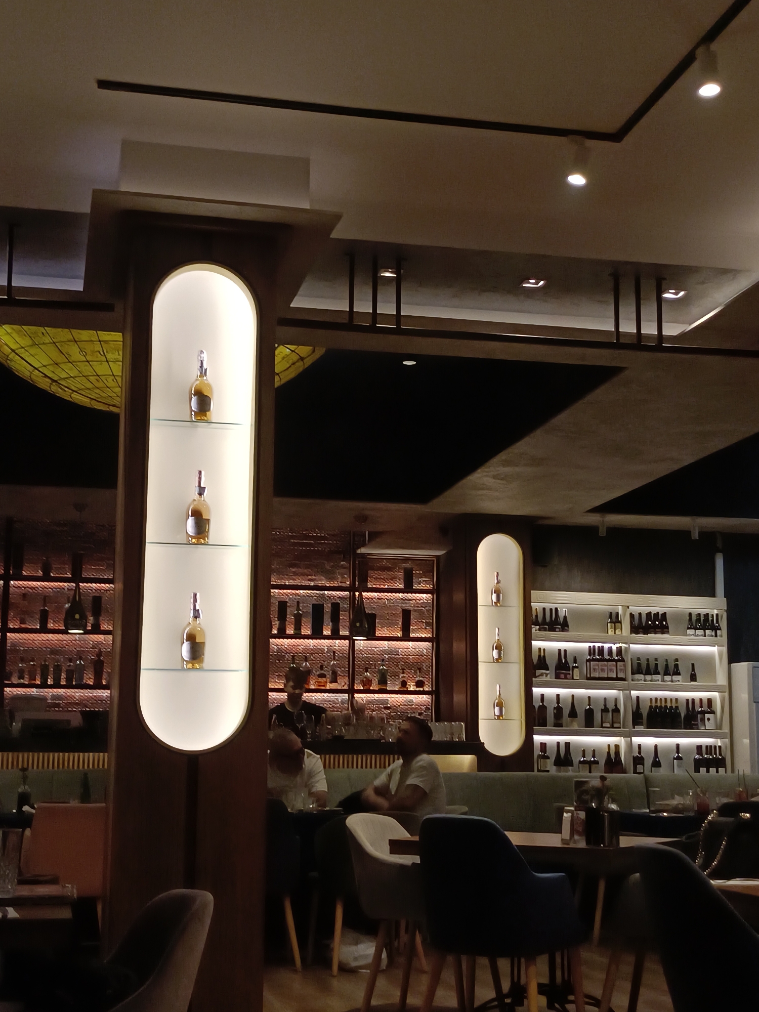 The image depicts the interior of a dimly lit bar or restaurant. The location is believed to be in Plovdiv, Bulgaria. Two illuminated vertical display cases are visible, showcasing three bottles each. Behind the display cases, shelves lined with bottles of liquor and wine create a backdrop. Patrons are seated at tables, some interacting with a bartender. The room features a modern ceiling with a grid-like design and recessed lighting. Dark upholstered chairs and booths are arranged around the tables.