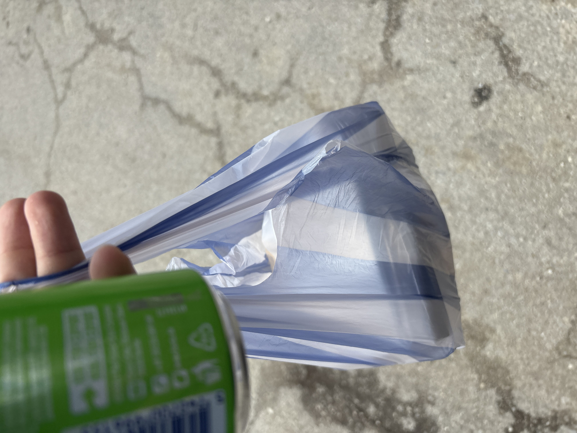 A hand holds a green can inside a striped plastic bag. The background shows a concrete surface with cracks. The location is unknown.