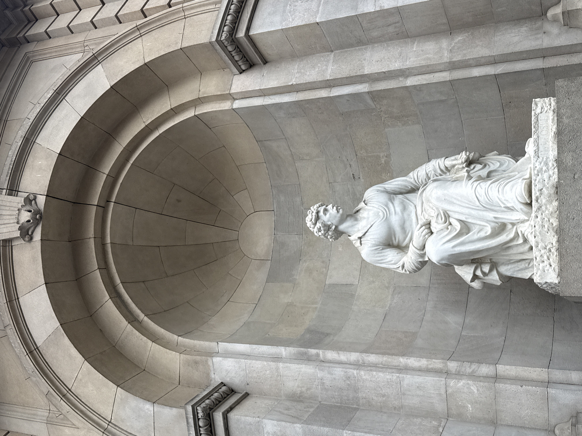 A marble statue of a reclining male figure is set within an arched alcove in Barcelona, Spain. The alcove, constructed of grey stone, features a series of concentric arches that lead to a dome-shaped interior. The statue is positioned on a ledge within the alcove. The figure is dressed in draped clothing, with his head and upper body visible. A stone plaque is set on the right edge of the alcove, adjacent to the statue. The stonework exhibits signs of weathering and age.