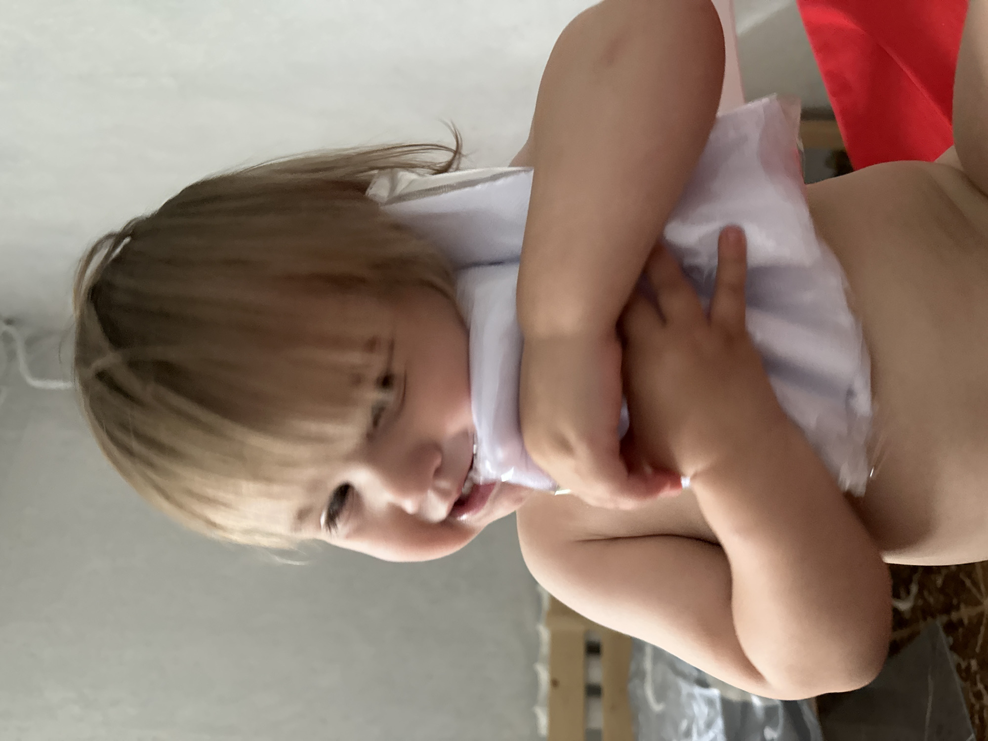 A child with blonde hair is holding a white object to their face. The child is looking to the left and smiling. The child's arms are around the white object, and their hands are visible. The child appears to be indoors, in a room with a bed in the background. The lighting suggests it is daytime. The location is not identifiable from the image alone.