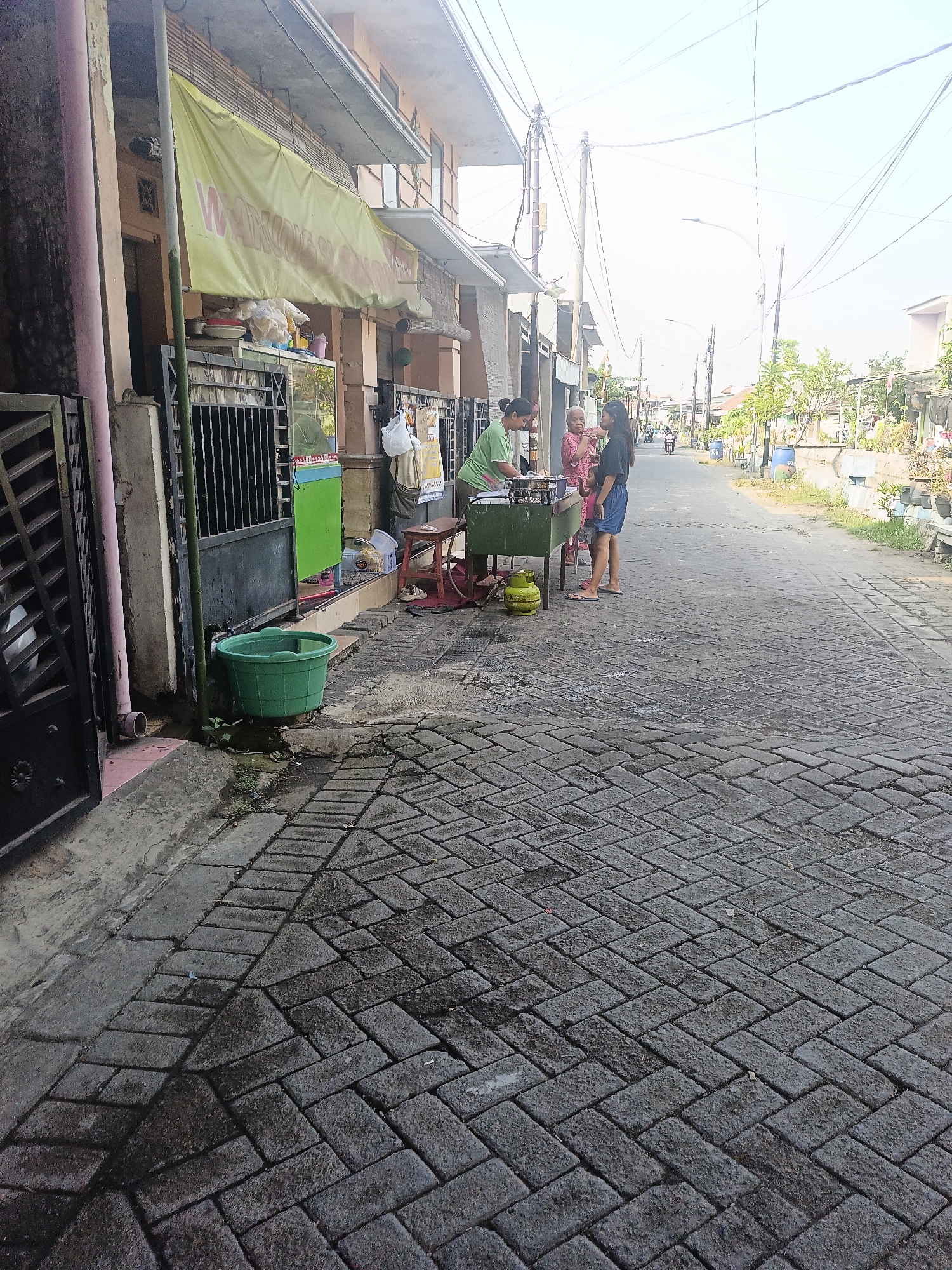In the city of Gedangan, Indonesia, a street scene unfolds with a modest food stall situated on the right side of the image. The stall, characterized by a green metal counter and a visible yellow propane tank, is attended by three individuals. A woman in a green shirt appears to be preparing food, while an older woman in a pink patterned shirt and a younger woman in a black shirt observe. The foreground is dominated by a paved walkway constructed from dark gray interlocking bricks, exhibiting some wet patches and wear. To the left, a dark metal gate and a portion of a building with a faded yellow banner adorn the facade. Beyond the stall, the street continues, lined with buildings and vegetation, with a clear view of electrical wires and poles stretching across the sky. The overall ambiance suggests a typical day in a residential area of Gedangan, with daily life activity centered around the small food establishment.