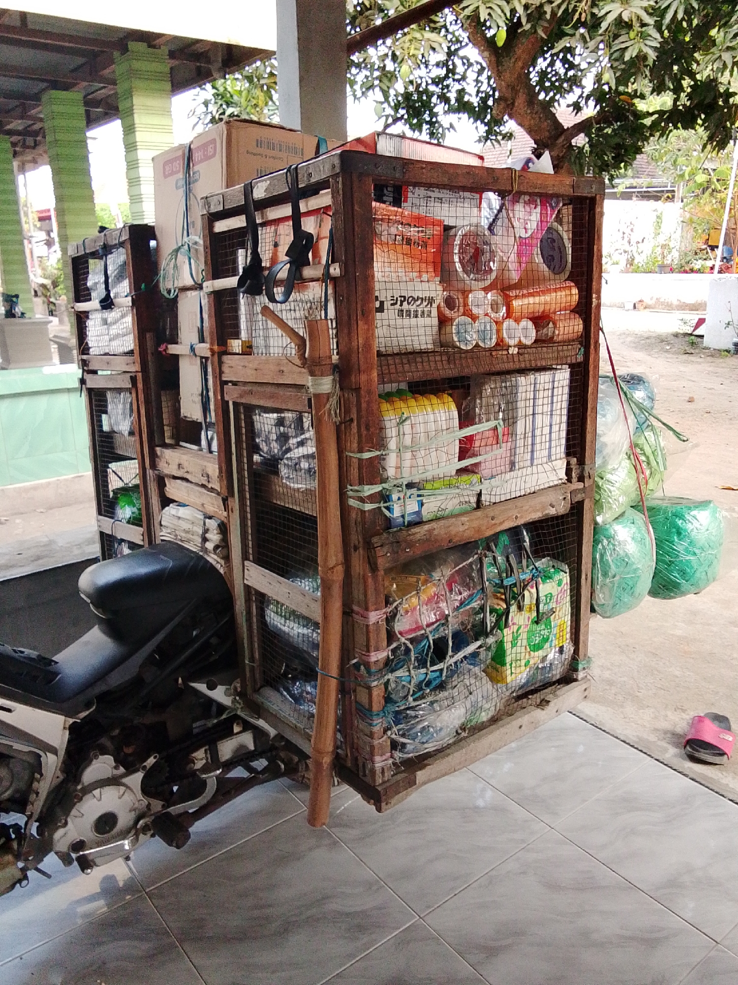 The image depicts a modified motorcycle with a large wooden cargo rack attached to the rear, laden with various goods. The motorcycle is parked on a tiled surface, likely a patio or sidewalk, in what appears to be an outdoor, possibly residential or commercial, area. Behind the motorcycle, there are green pillars supporting a roof structure. Further in the background, trees and buildings are visible. The cargo rack is constructed from rough-hewn wooden planks and wire mesh, with multiple shelves holding an assortment of items. These items include cardboard boxes, rolls of tape, plastic-wrapped bundles, and fabric bags. Some of the items are secured with ropes and bungee cords. To the right of the motorcycle, a single pink flip-flop is visible on the ground. The scene suggests a delivery or transportation operation, possibly in a town like Bulurejo, Indonesia, given the context. The lighting indicates it is daytime.
