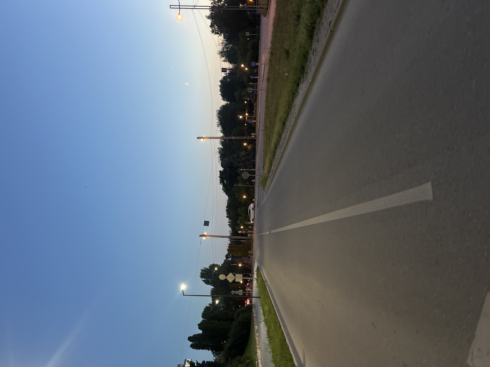 The image depicts a twilight scene on a multi-lane road in Ternopil, Ukraine. The sky is a deep blue, transitioning to a lighter hue near the horizon, with a faint crescent moon visible. Streetlights illuminate the road and surrounding areas.
On the left side of the road, there is a grassy embankment with trees and bushes. A few streetlights are present, and a white car is parked on the side. Further down the road, a group of people are gathered near some street signs.
The road itself is asphalt and appears to be in good condition, with a white line dividing the lanes. Several cars are visible driving in the distance. On the right side of the road, there is more vegetation and more streetlights. Traffic signs are scattered throughout the scene, indicating road directions and regulations. The overall atmosphere is calm and peaceful, suggesting a typical evening in the city.