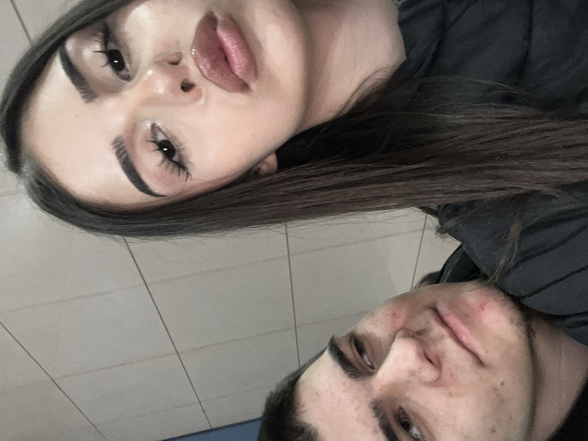 The image is a selfie taken at an upward angle, featuring two individuals. The first person is a female with dark hair, prominent eyebrows, and full lips. She appears to be closer to the camera. The second person is a male with short dark hair and light skin, positioned behind the female. The background consists of light-colored, square tiles, possibly a wall or ceiling. The overall scene suggests an indoor location. There is no information available in the image to determine the location in the city of Sofia, Bulgaria.