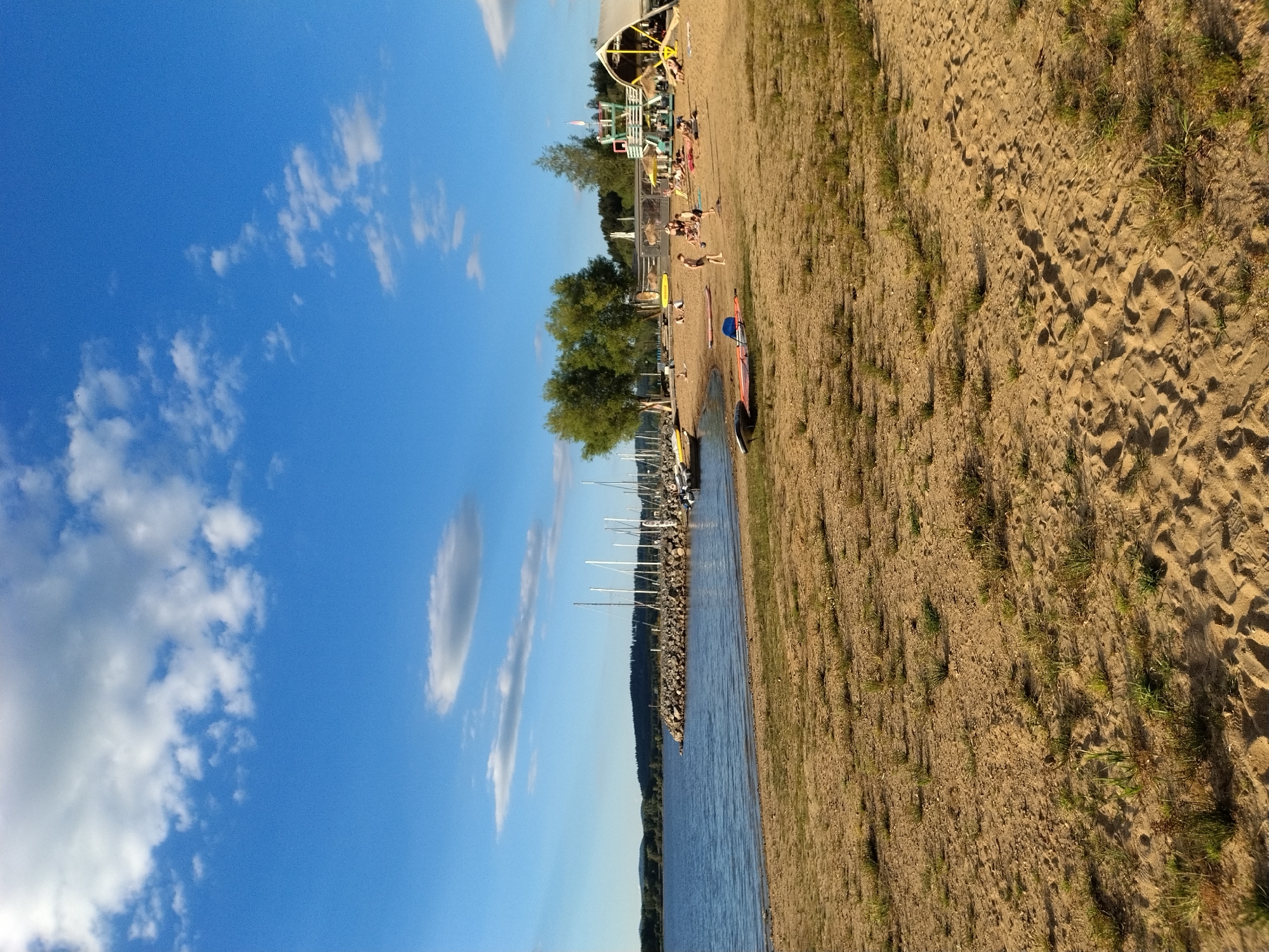 The image depicts a sandy beach on a sunny day, with a calm body of water to the left and a clear blue sky with scattered clouds above. In the background, a rocky breakwater separates the beach from a body of water where several sailboats are moored. Trees and hills form the distant shoreline.
On the beach, a small group of people are engaged in various activities. Several individuals are lying on beach towels, relaxing. Some children are playing in the sand, one appears to be kicking a ball, while others are seated or standing. There is a structure that resembles a lifeguard tower or a small observation deck with a yellow slide. Further to the right, a large tent-like structure with a thatched roof is partially visible. A few surfboards or paddleboards are lying on the sand. The overall scene suggests a recreational area, possibly a beach resort or a public park with a lake. The mention of Radslav, Czechia, places this scene in that specific geographical location, implying it is a popular spot for locals and tourists to enjoy outdoor activities during warmer weather.
