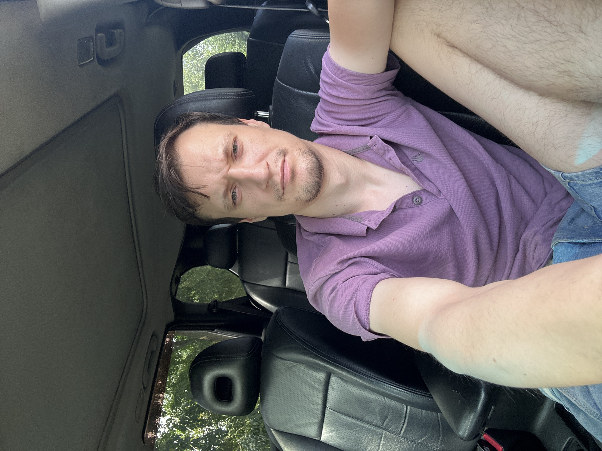 A man is seated in the driver's seat of a car, looking directly at the camera. He is wearing a short-sleeved, purple polo shirt and denim shorts. The interior of the car features black leather seats and a sunroof, visible in the upper portion of the image. Trees and foliage are visible through the car's windows, suggesting an outdoor setting. The image does not contain information indicating the location in the city of Makiv, Ukraine.