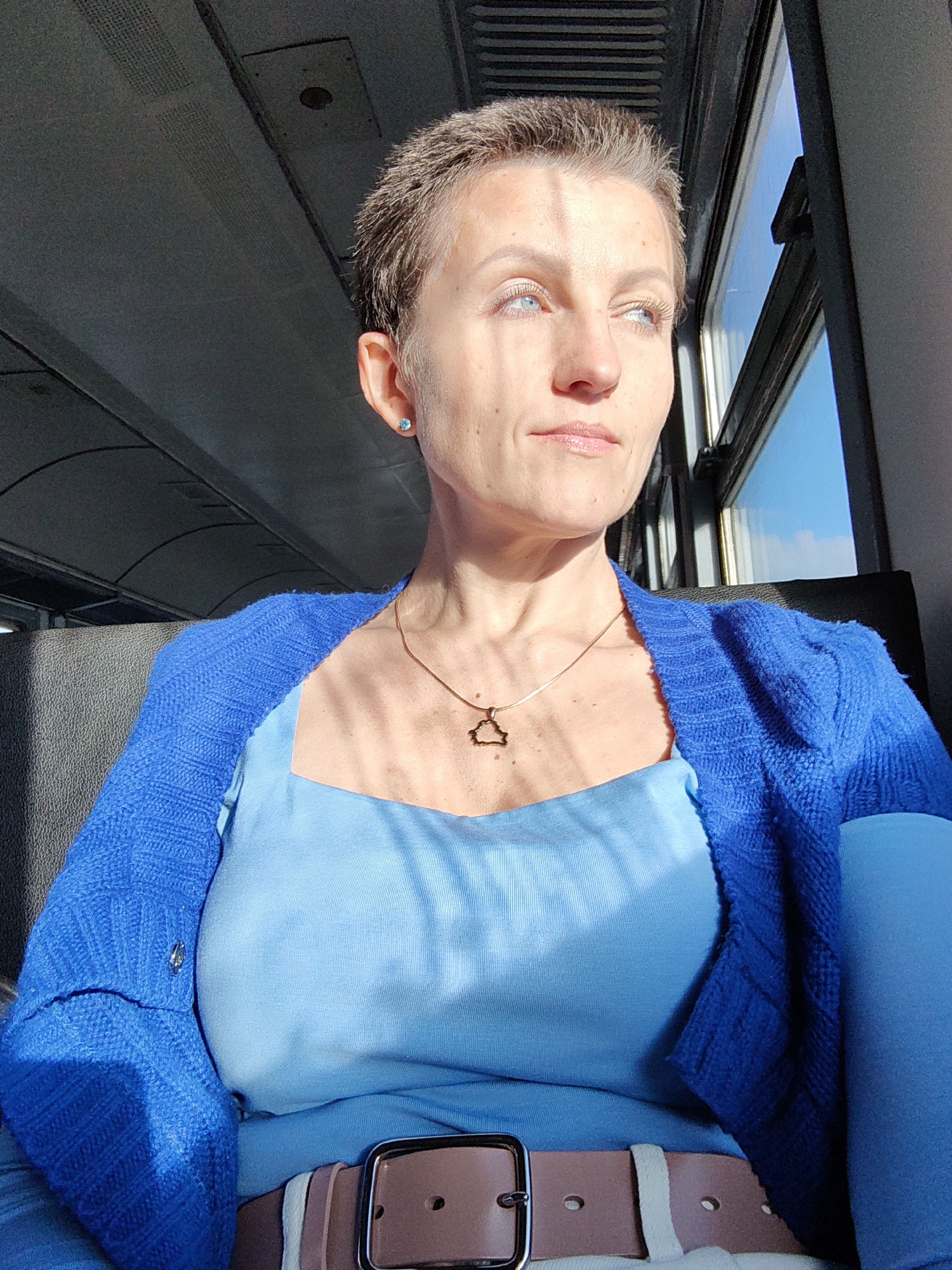 A close-up shot shows a woman with short, tousled brown hair and light blue eyes, looking to the right, bathed in sunlight that casts shadows on her face. She is wearing a light blue scoop-neck top and a bright blue knit cardigan. A delicate gold chain with a triangle pendant rests on her collarbone. A silver earring with a blue stone is visible on her left earlobe. She is seated, with a dark gray seat behind her and a window on the right revealing a clear blue sky. The environment appears to be inside a vehicle, possibly a train, given the textured ceiling and the window design. The woman is wearing a pale pink belt with a silver buckle. The overall color palette is dominated by blues and grays, with the sunlight adding warmth and contrast.