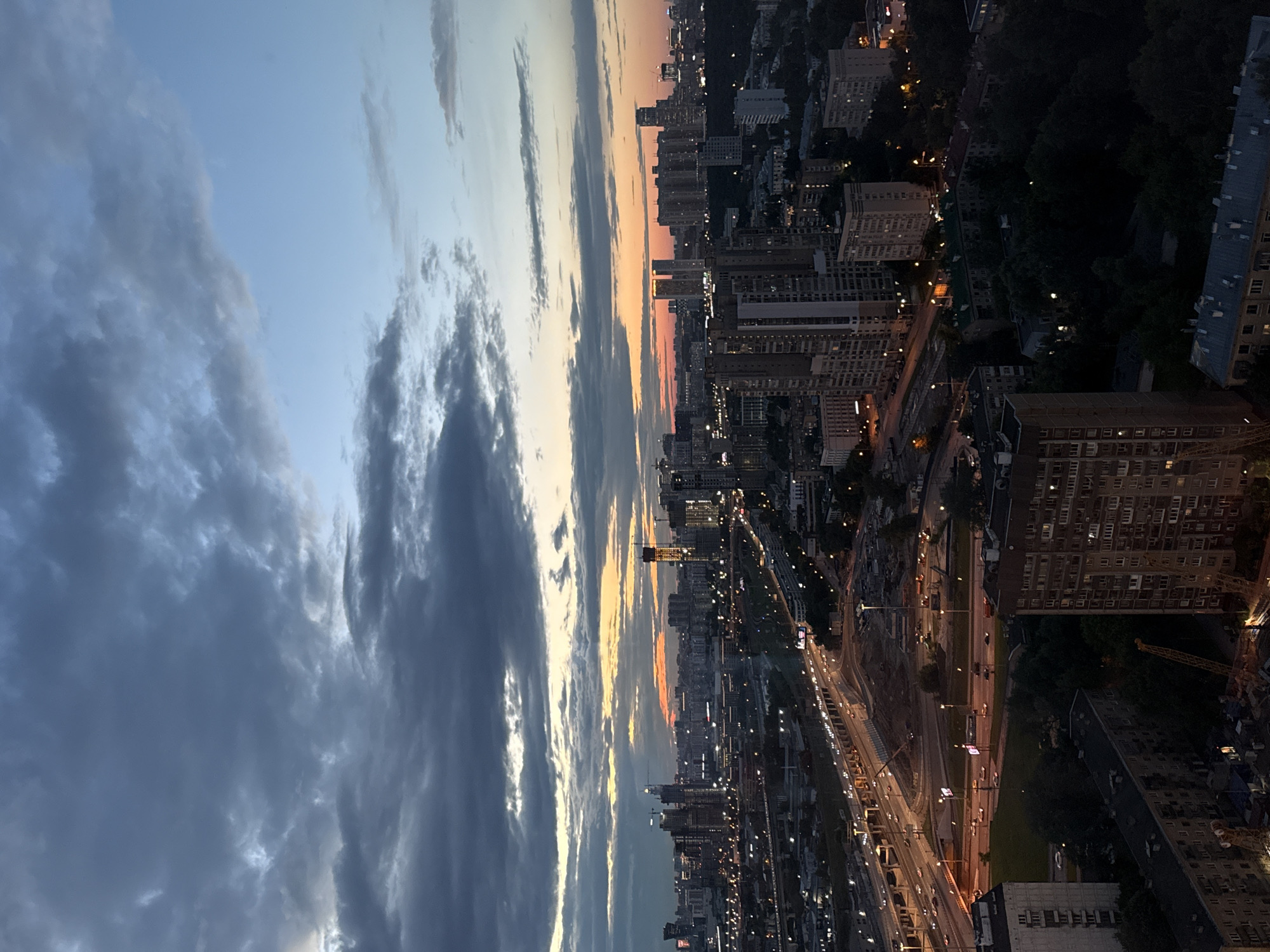 The image captures a nighttime cityscape in Khimki, Russia, with a dramatic sky and a sprawling urban landscape. The photo is oriented vertically, with the sky occupying the left half and the city stretching across the right. The sky transitions from a pale blue at the top to hues of orange and pink near the horizon, suggesting either sunrise or sunset. Dark, wispy clouds are scattered across the upper portion of the sky, adding texture and depth.
Below the sky, the city is illuminated by a multitude of lights. Numerous high-rise buildings, characteristic of urban Russia, dominate the scene. Their windows glow with warm light, creating a grid-like pattern against the darkening sky. A busy highway runs through the city, with streaks of headlights and taillights indicating a steady flow of traffic. Streetlights and lights from various structures further illuminate the urban sprawl.
Notable details include construction cranes, hinting at ongoing development within the city. The overall atmosphere is one of quiet activity, typical of a city at dusk or after dark. The scene evokes a sense of scale and the vastness of the urban environment. No visible text is present in the image.