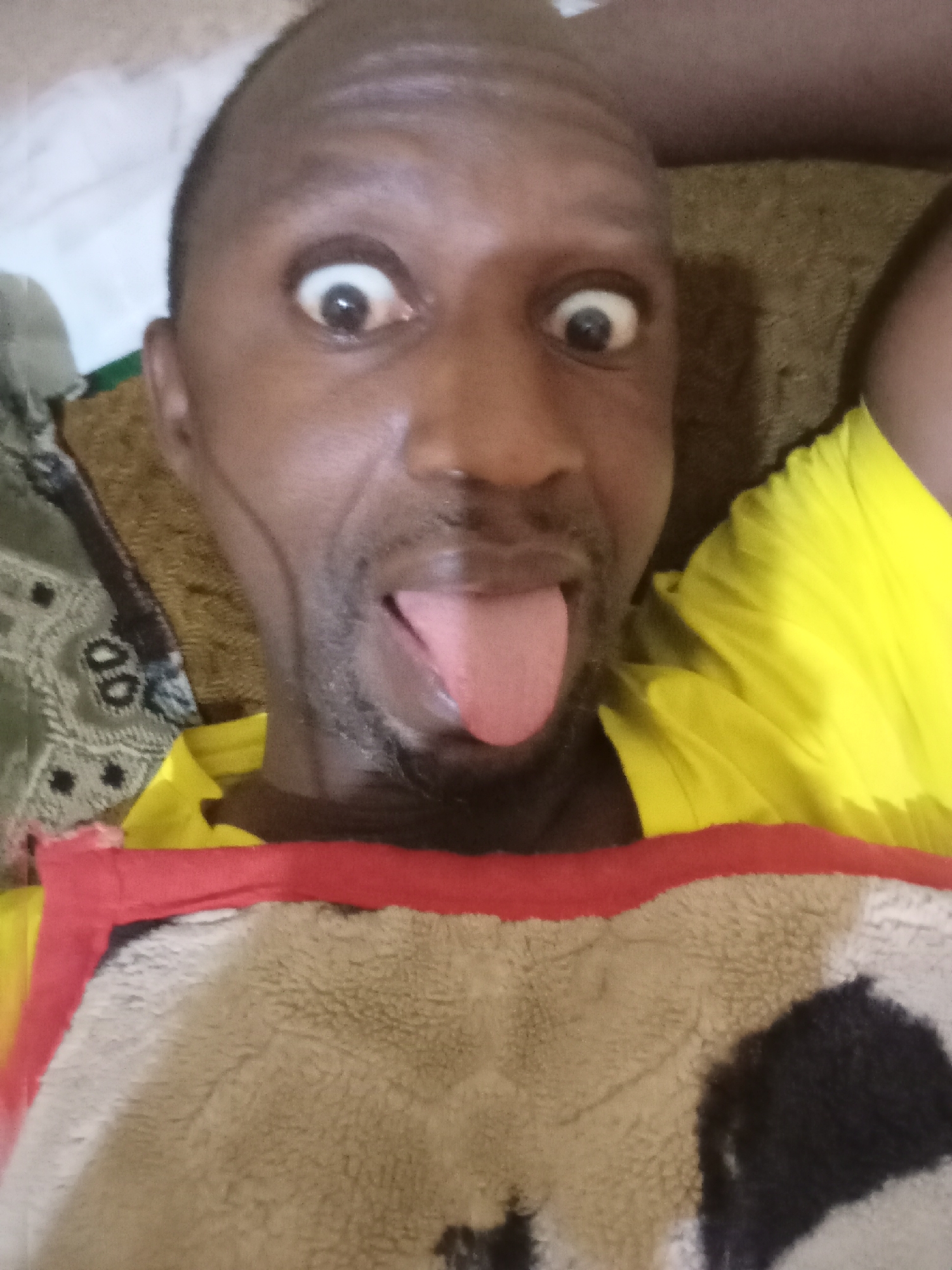 A close-up selfie depicts a dark-skinned man with a shaved head making a face. His eyes are wide open, and he is sticking out his tongue. He is wearing a yellow shirt or garment, and a patterned blanket with red trim is partially visible below his neck. The background is obscured, showing only a bed and bedding in neutral tones. The location is not discernible as Jos, Nigeria, from the visual information provided.