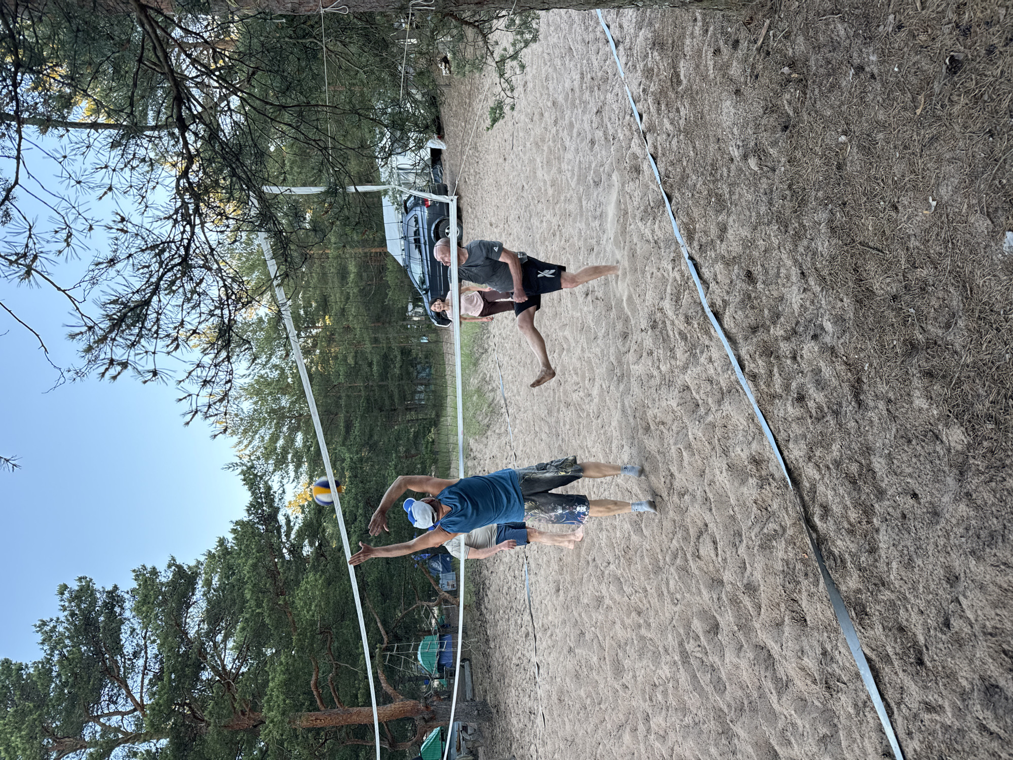 In Ozerki (Krasnodolinskaya), Russia, four individuals are engaged in a game of beach volleyball on a sandy court surrounded by pine trees. A volleyball is suspended in the air above the net, indicating an active play. Two men are on one side of the net, one in a blue tank top and shorts appears to be mid-jump, reaching for the ball, while another man in a grey t-shirt and black shorts is kicking the ball. On the other side of the net, a woman in a pink t-shirt and dark pants and a man in a light-colored t-shirt and blue shorts are positioned on the court. In the background, tents and a car suggest a campground or recreational area. The sky is clear and blue.