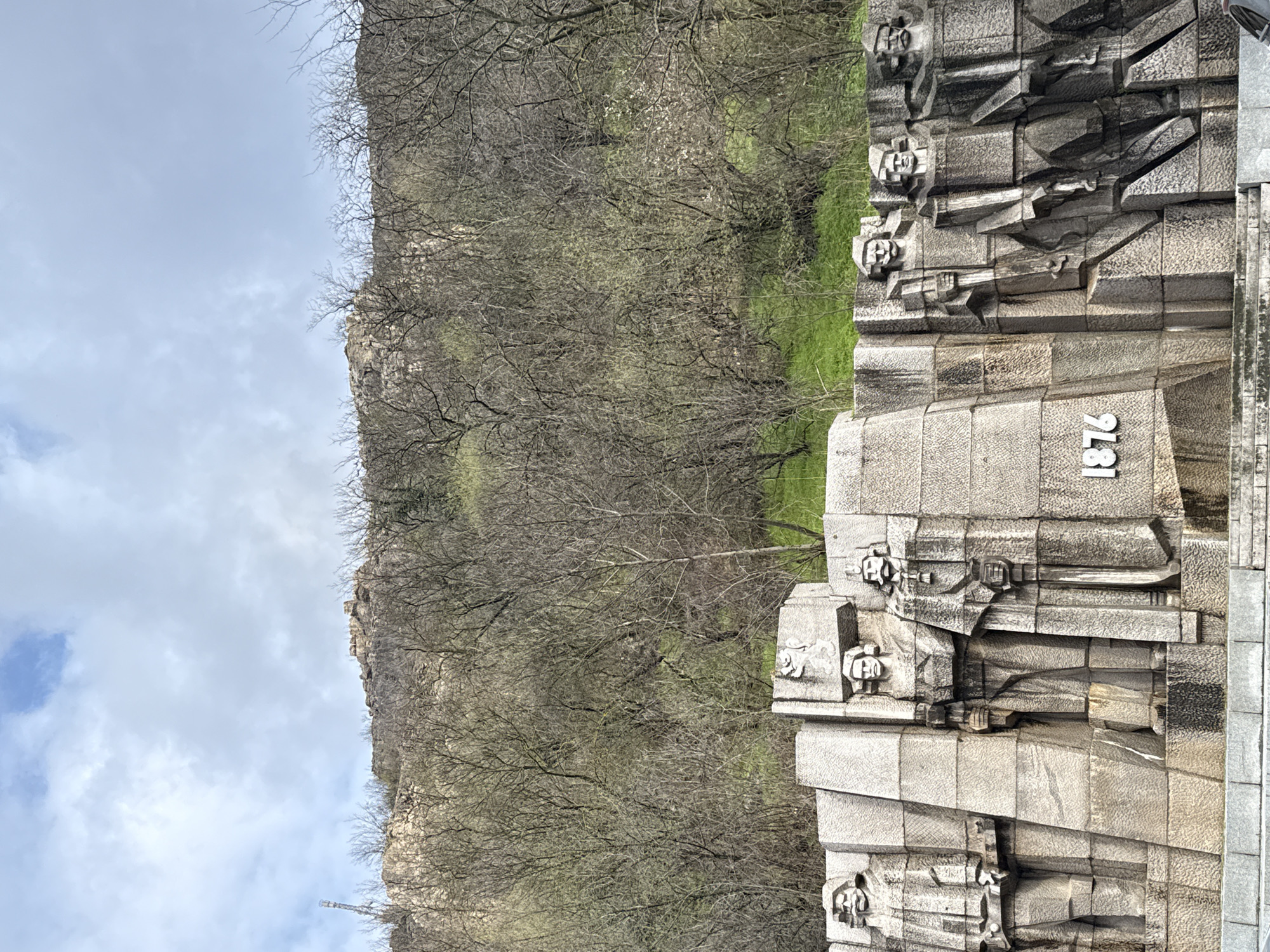 The image shows the Monument to Vasil Levski in Lovech, Bulgaria, set against the backdrop of a rocky hillside with sparse vegetation. The monument is constructed of light gray stone blocks and features bas-relief depictions of human figures, seemingly soldiers. The inscription "1876" is prominently displayed on one of the blocks. The background includes a hill covered in trees without leaves, and a sky with scattered clouds.