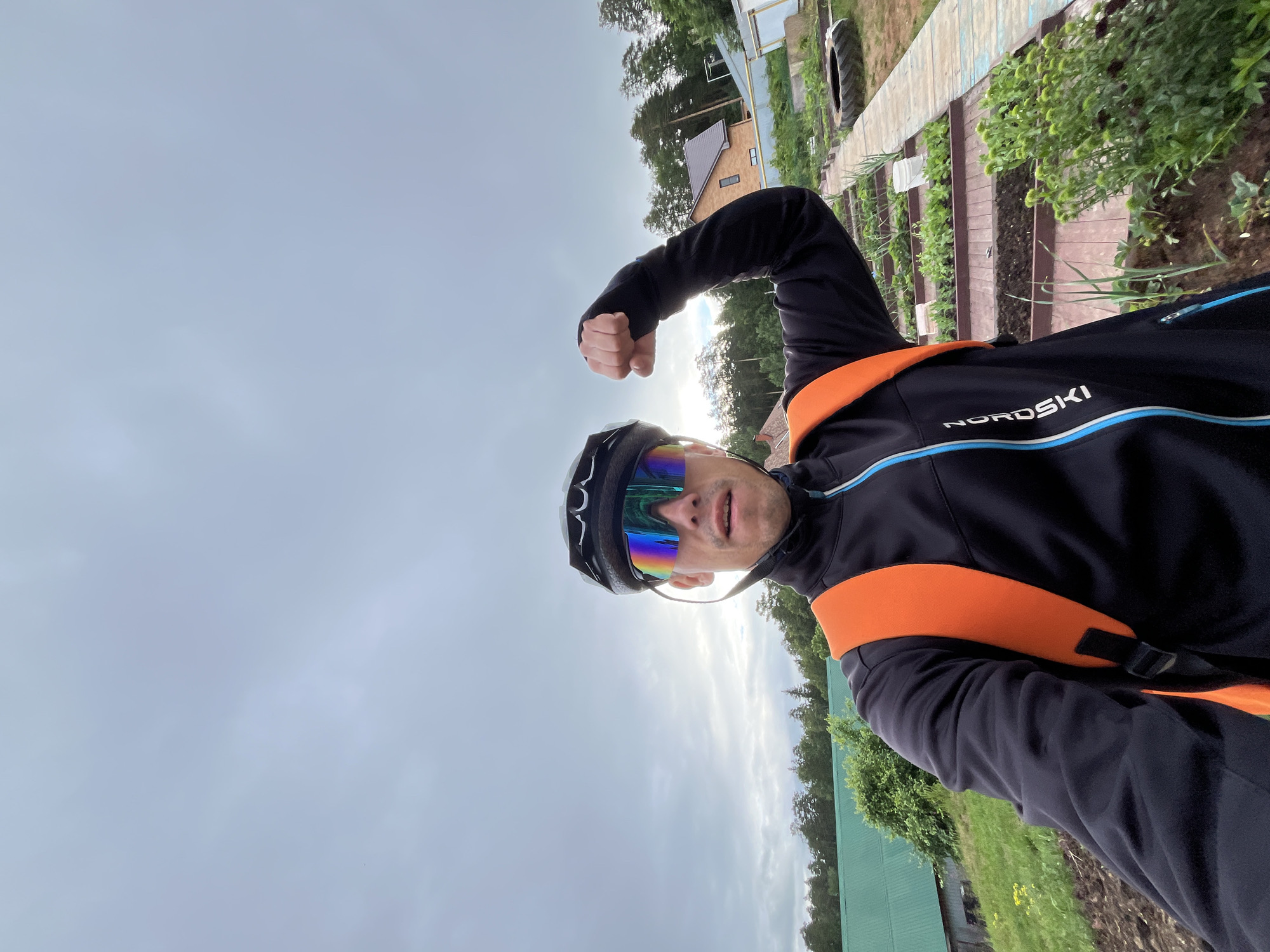 A man wearing a black cycling helmet, iridescent cycling glasses, and a black jacket with an orange backpack is posing with his right fist raised in a flexed bicep position. He is standing in what appears to be a backyard or garden area. In the background, there are raised garden beds with young plants, a wooden walkway, trees, and a house with a red-tiled roof. The sky is overcast. The image was taken in the city of Chaikovsky, Russia.