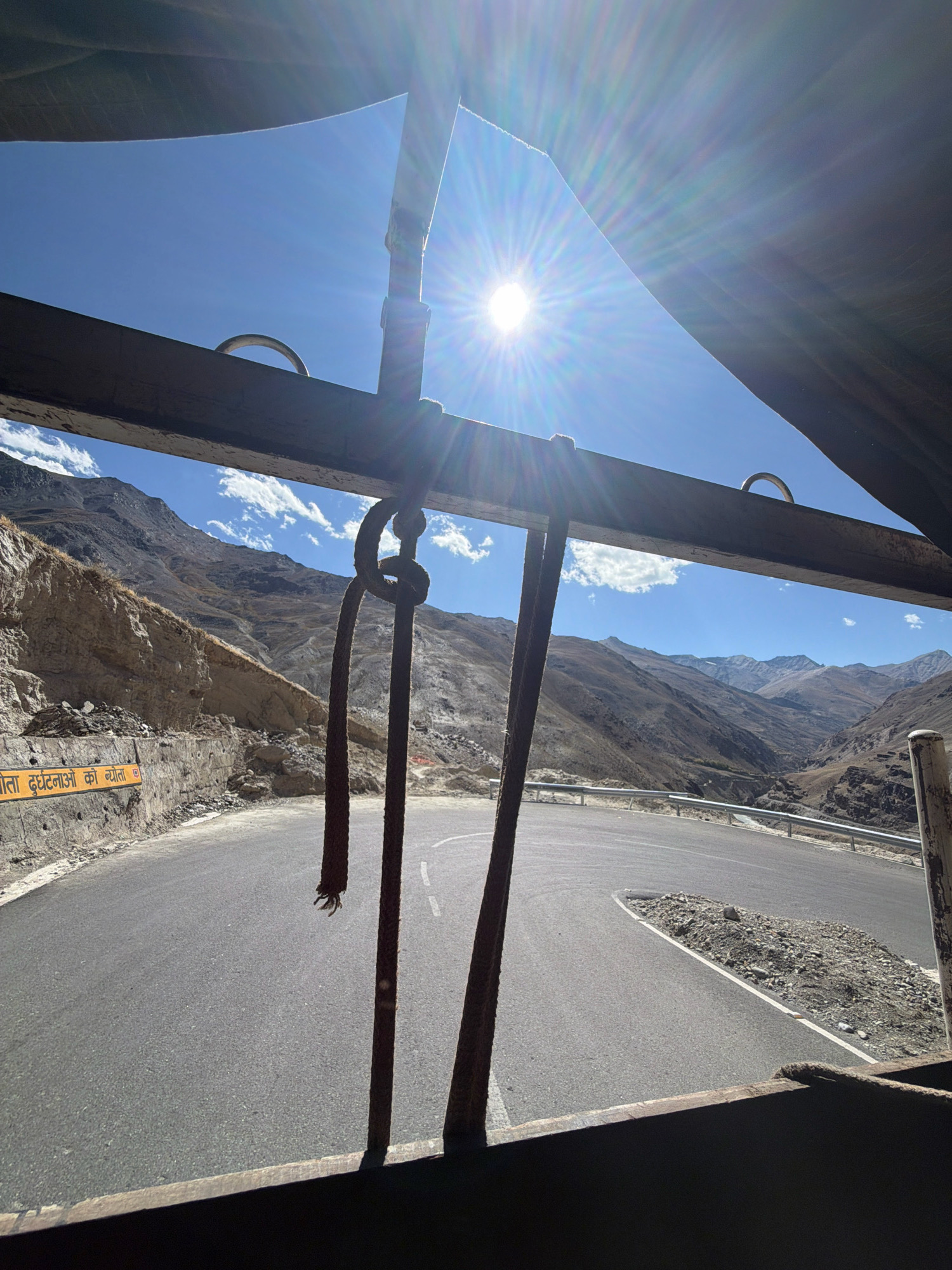The image depicts a winding mountain road in a rural, arid environment, likely in the region of Char, India. A bright sun shines high in a clear blue sky, creating lens flares. In the foreground, a wooden beam runs horizontally across the frame, with two thick ropes hanging from it. The road is paved with asphalt and has a white dashed line marking the center. To the left of the road, a yellow sign with Hindi text is mounted on a concrete wall. The landscape is characterized by dry, rocky mountains that rise steeply on either side of the valley. The overall scene suggests a journey through a remote and mountainous terrain under a sunny sky.