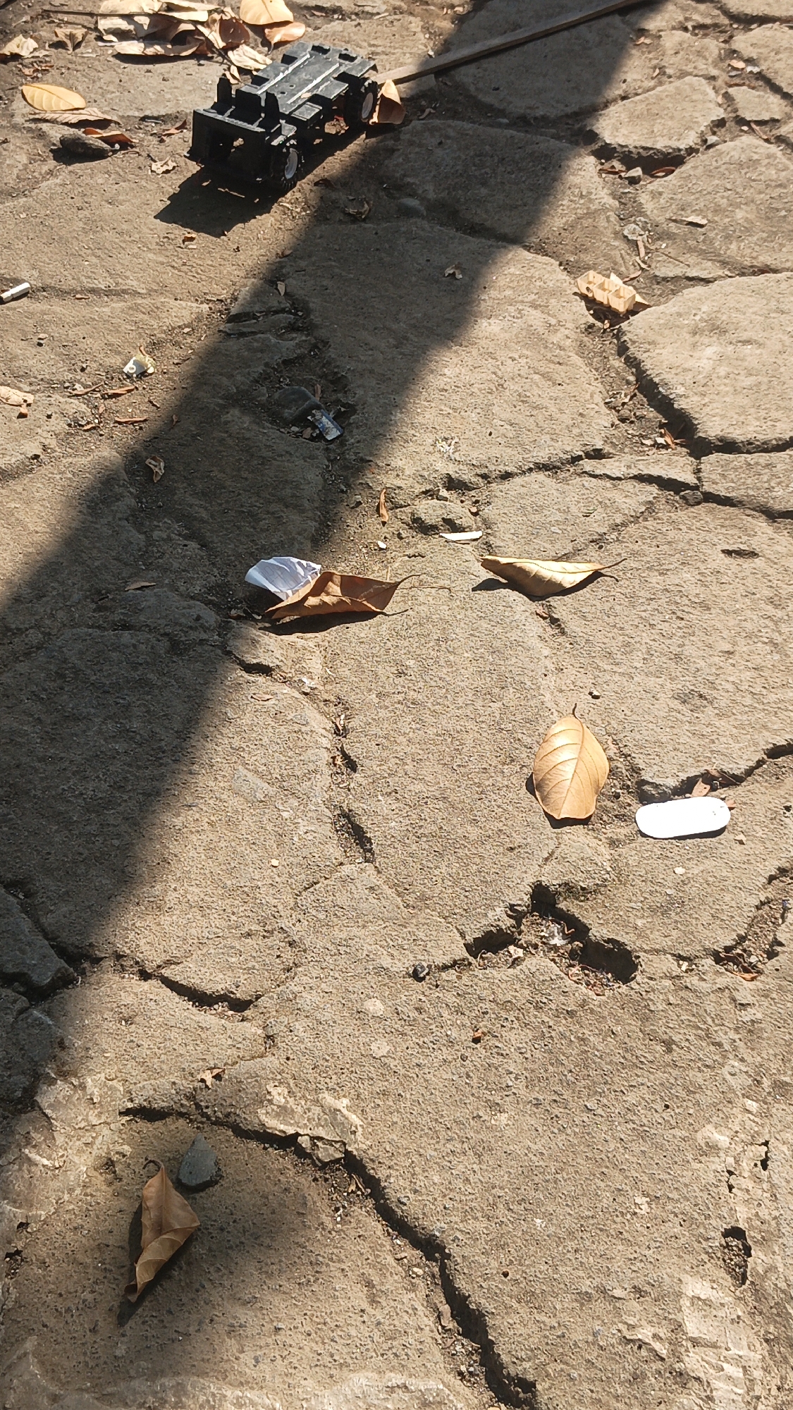 A toy truck and scattered dry leaves rest on a cracked concrete surface. The scene is lit by sunlight, casting a long shadow across the ground. There is some debris present, including a small white object and what appears to be a discarded cigarette butt. The location is identified as Bulungcangkring, Indonesia.
