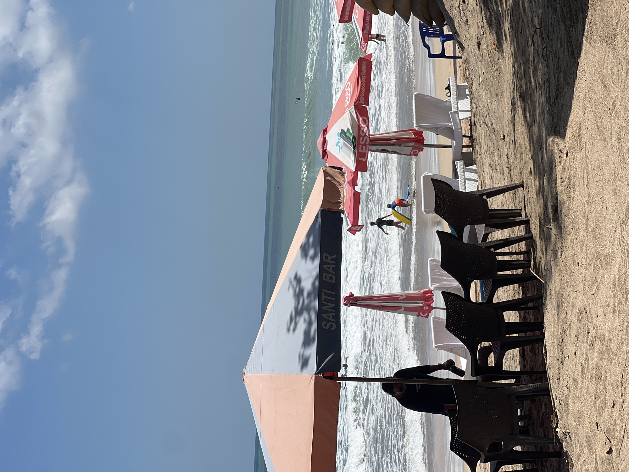 The image depicts a sunny day at Kuta Beach, Bali, Indonesia, a popular tourist destination known for its surfing. In the foreground, several dark plastic chairs are arranged around tables on the sand, suggesting a seating area for a beachfront establishment. One large, two-toned umbrella, orange and gray, is prominently featured, with the words "SANTI BAR" visible on its side. To the right, two red and white umbrellas, one with "ESSO" and "LESSO" branding, are set up closer to the water.
The middle ground shows a stretch of wet sand leading to the ocean. Several beach chairs, appearing to be white loungers, are placed near the water's edge. In the sea, there are several individuals. Some are surfing, with one person clearly visible riding a wave. Others appear to be wading in the water, including a child in an orange shirt holding a yellow floatation device.
The background consists of the vast blue ocean with moderate waves, indicating a suitable condition for surfing. The sky is clear blue with a few scattered white clouds. The overall atmosphere is that of a lively beach scene, with people enjoying water activities and the amenities provided by beachside businesses. The presence of the "SANTI BAR" umbrella suggests this is a commercial area within Kuta.