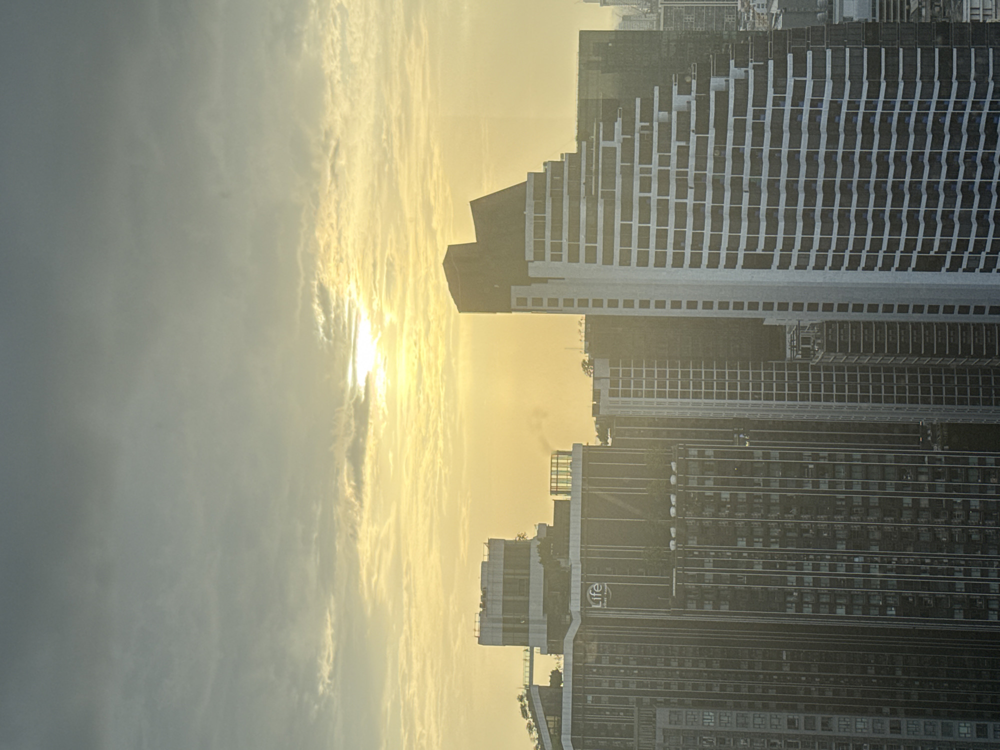 This is a high-angle, outdoor, daylight, vertical photograph of a cityscape during sunrise or sunset. The sun is partially obscured by clouds, casting a warm, golden glow over the scene. Multiple high-rise buildings dominate the view, with a particularly prominent, uniquely shaped skyscraper in the foreground to the right, featuring a stepped facade with alternating dark and white sections. To the left, another large building displays the "Life" logo. The sky is filled with textured clouds, tinged with yellow and orange hues. The overall impression is one of urban development against a natural atmospheric phenomenon.
