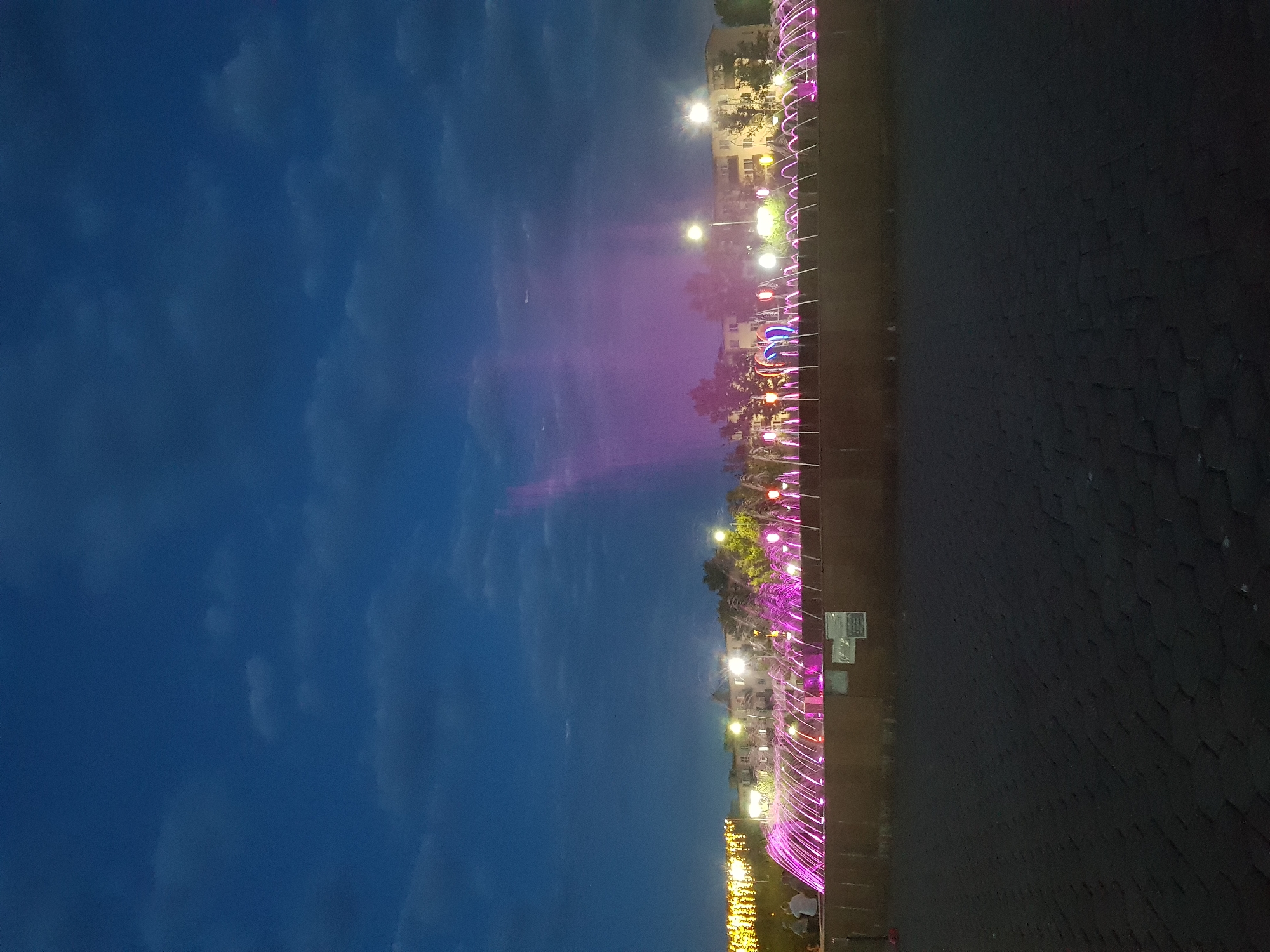 An outdoor, low-angle, eye-level, long shot shows a public fountain at dusk in Kurchatov, Kazakhstan. The fountain is illuminated with pink and purple lights, with streams of water arcing upwards. Behind the fountain, there are trees and buildings with lights on. The sky is a deep blue with scattered clouds. In the foreground, a paved walkway is visible. There are a few silhouetted figures of people standing near the fountain.