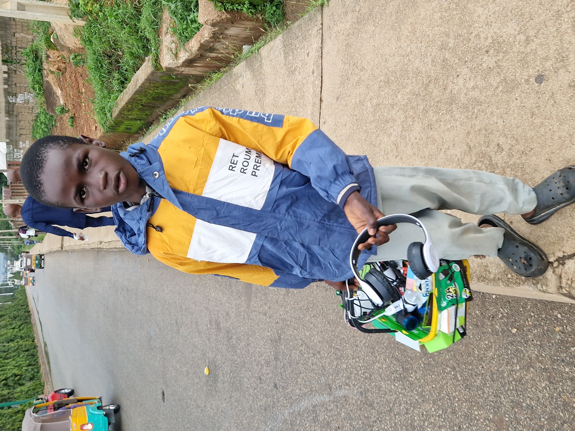 In Jos, Nigeria, a young boy stands on a sidewalk next to a road, holding a basket filled with various items, including a pair of headphones. He is wearing a blue and yellow jacket, light-colored pants, and black crocs. The boy is looking directly at the camera. In the background, other people and vehicles are visible, including a colorful auto-rickshaw and a person in a blue shirt walking away from the camera. The environment includes a grassy embankment and a concrete wall.