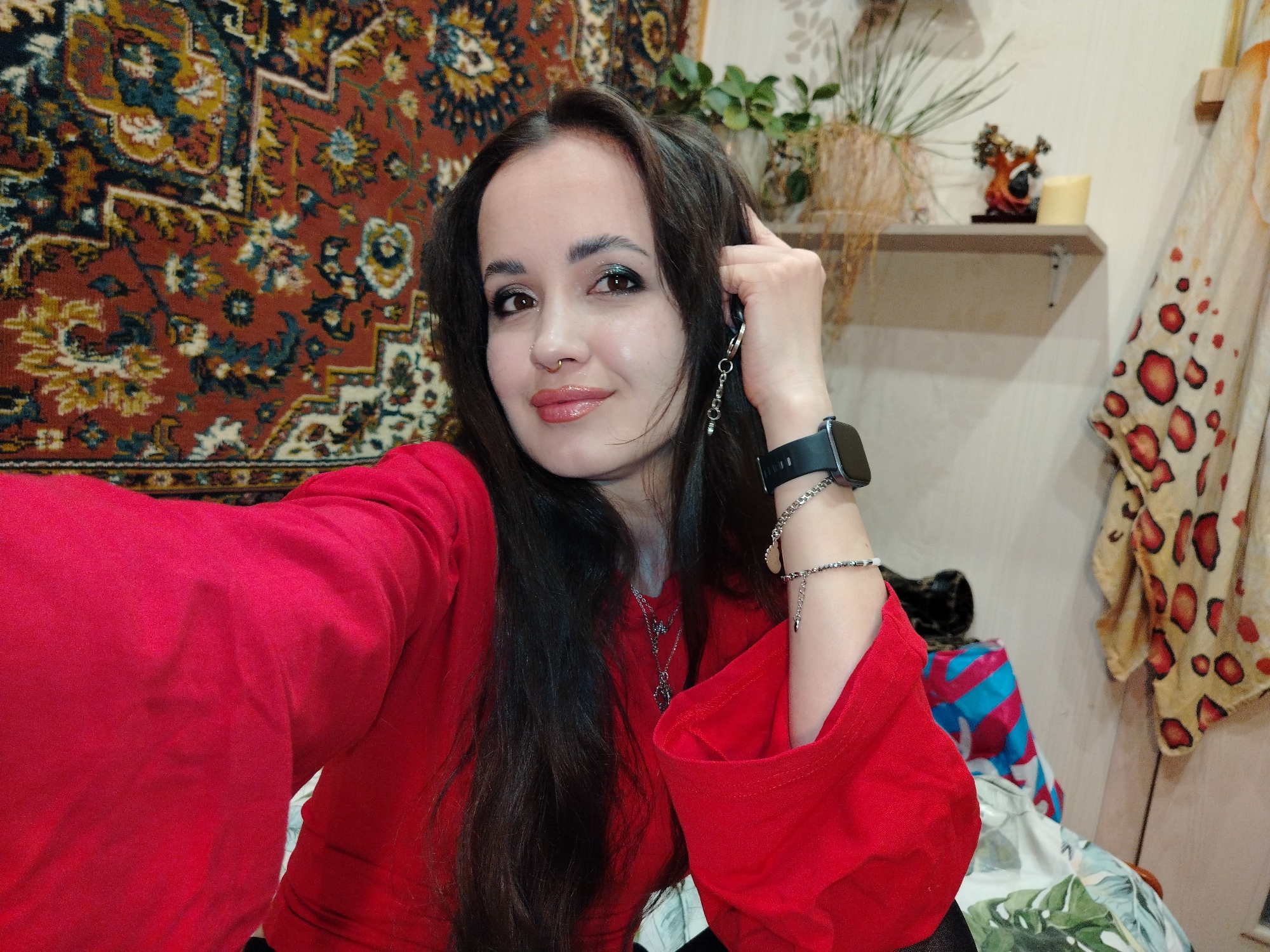 The scene is set indoors in a domestic environment in Unknown, Unknown. A single female subject with long, dark hair, dark eyes, and a slight smile is depicted in a frontal, close-up view. She wears a red long-sleeved top, a gold-toned nose ring in her right nostril, green eyeliner, and pink lip gloss. A black smartwatch and a silver-colored bracelet with charms are visible on her left wrist. She also wears a silver-colored necklace with multiple pendants. Her left hand is raised, with fingers near her temple, holding a silver-colored keychain bearing a "Love" charm.
The background to the subject's left features a large tapestry or rug displaying an intricate floral and geometric pattern in red, blue, yellow, and cream tones. To the subject's right, a light-colored wall is visible. A wooden shelf on the wall supports a potted plant, a small decorative figurine in orange and brown tones, and a cream-colored cylindrical object. Below the shelf and to its right, a light-colored fabric with red circular patterns hangs from a wooden peg. Additional patterned fabrics are partially visible in the lower right foreground.