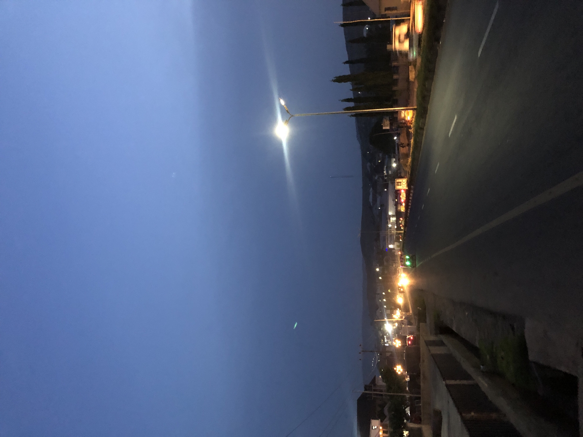 The image captures a roadside scene at dusk or night in Karu, Nigeria. The sky, occupying a significant portion of the left side, is a deep, clear blue, transitioning from lighter blue at the top to a darker hue towards the bottom, with a faint white dot visible in the upper left. A prominent streetlight with a bright, star-like glare illuminates the mid-ground, casting light across the scene.
On the right side, a wide, dark asphalt road stretches into the distance, marked with white dashed and solid lines. Alongside the road is a concrete barrier or drainage ditch, beyond which various buildings and structures are visible, their windows and signs emitting warm yellow and orange lights. Several slender, dark trees, possibly cypress, line the horizon, silhouetted against the sky and distant, dark hills or mountains.
No people or vehicles are directly visible, suggesting a quiet moment on the thoroughfare. The artificial lights from streetlights and establishments along the road are a notable feature, indicating the late hour. A small yellow sign with the text "SCC" in black letters is discernible on the left side of the road, hinting at a local business or infrastructure element. The overall atmosphere is serene, with the artificial lights contrasting with the natural darkness of the night and distant landscape.