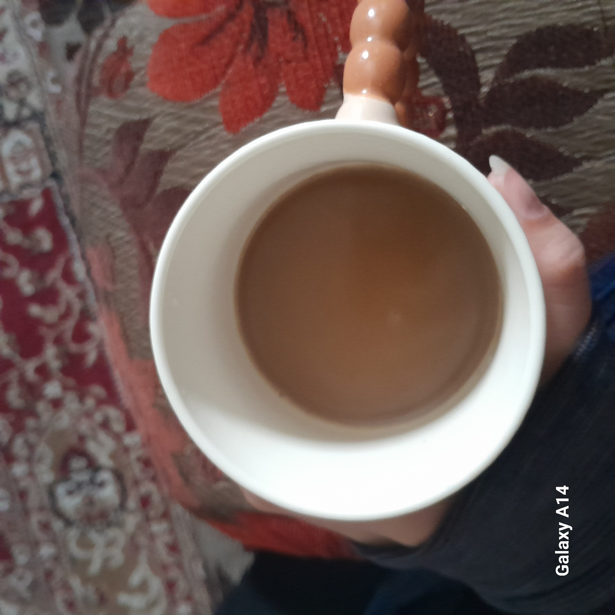A top-down shot shows a white mug filled with a brown liquid, presumably coffee or tea with milk. The mug is being held by a person with manicured nails. A portion of the person's grey-blue sleeve is visible. The mug is positioned over a surface with a floral pattern in shades of red and brown. The bottom right corner of the image includes the text "Galaxy A14." The location in the city is unknown.
