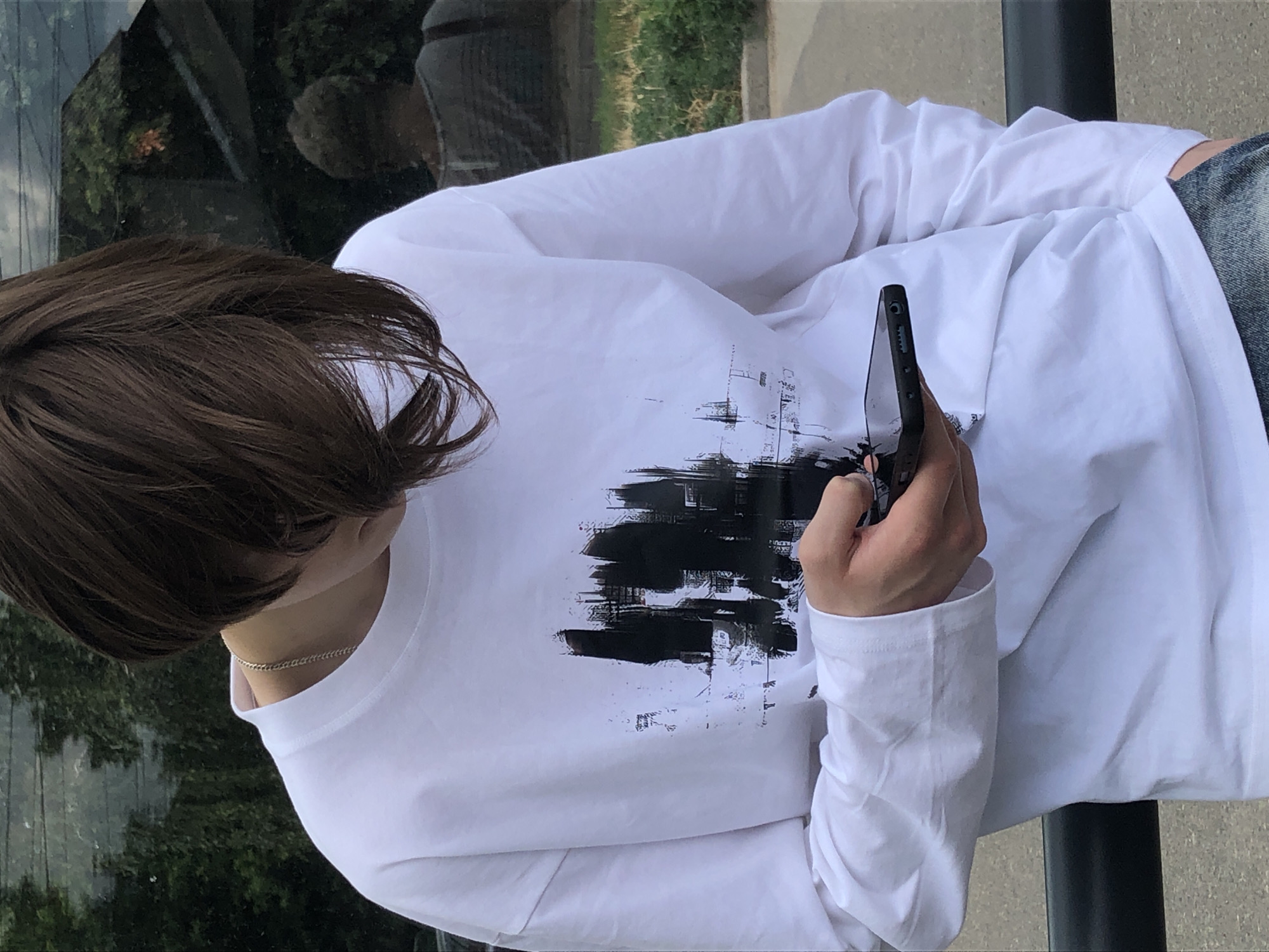 The image shows a person from the chest up, wearing a white long-sleeved shirt with a black graphic design on the front. The person has dark brown hair and is looking down at a black smartphone they are holding in their right hand. They are wearing a silver chain around their neck. In the background, there is greenery and a reflection in what appears to be a glass surface, showing another person and some trees. The scene suggests an outdoor urban or park setting. There is no specific information within the image that points to the city of Chernihiv, Ukraine.