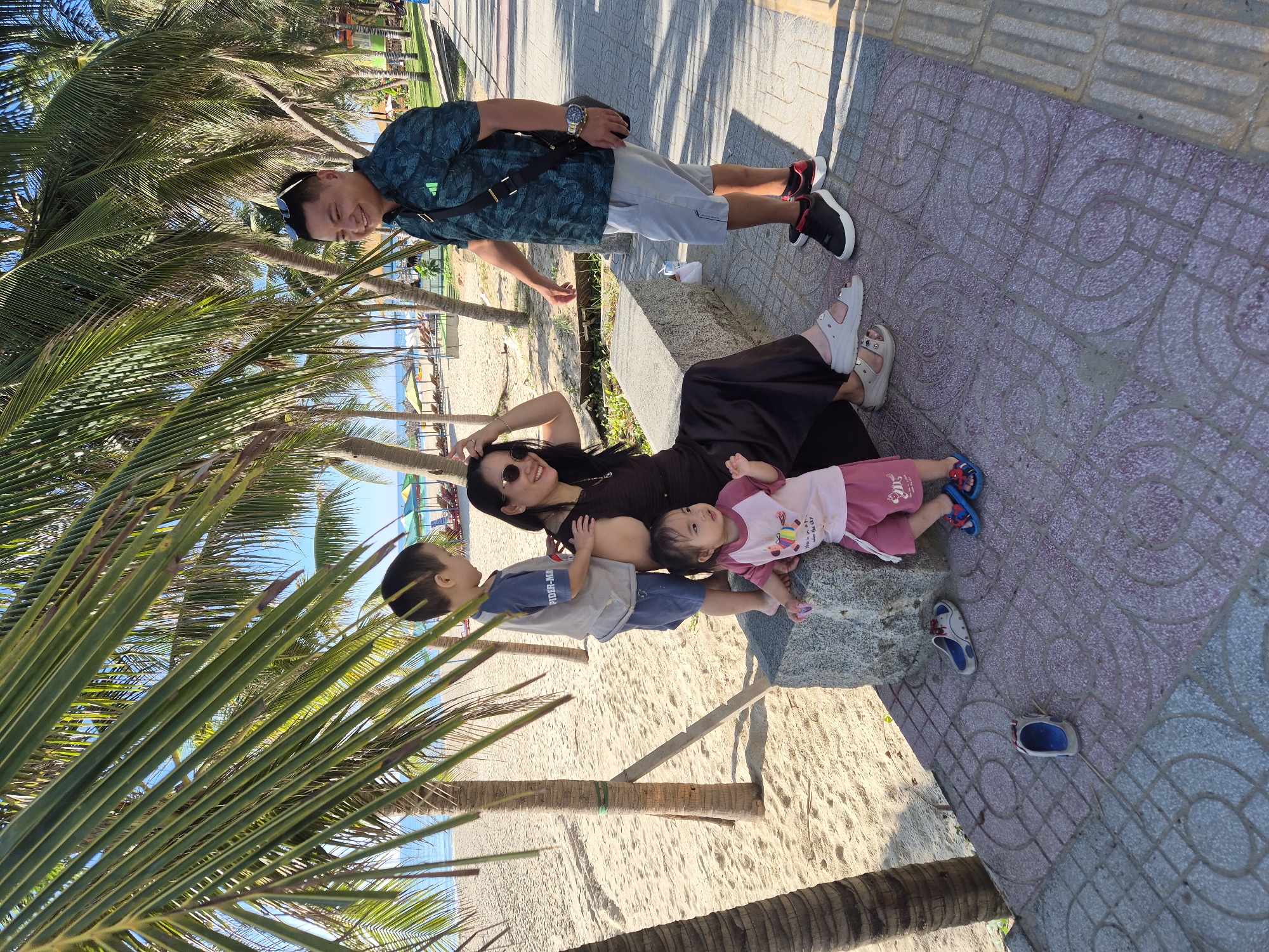 The image depicts a family enjoying a day at the beach in Da Nang, Vietnam. A man and woman are seated on a stone bench, with two young children standing near them. The beach is visible in the background, with palm trees lining the shore. The man is wearing a green patterned shirt and grey shorts, while the woman is in a dark dress. The children are dressed in casual clothing. The scene appears to be a relaxing family outing.