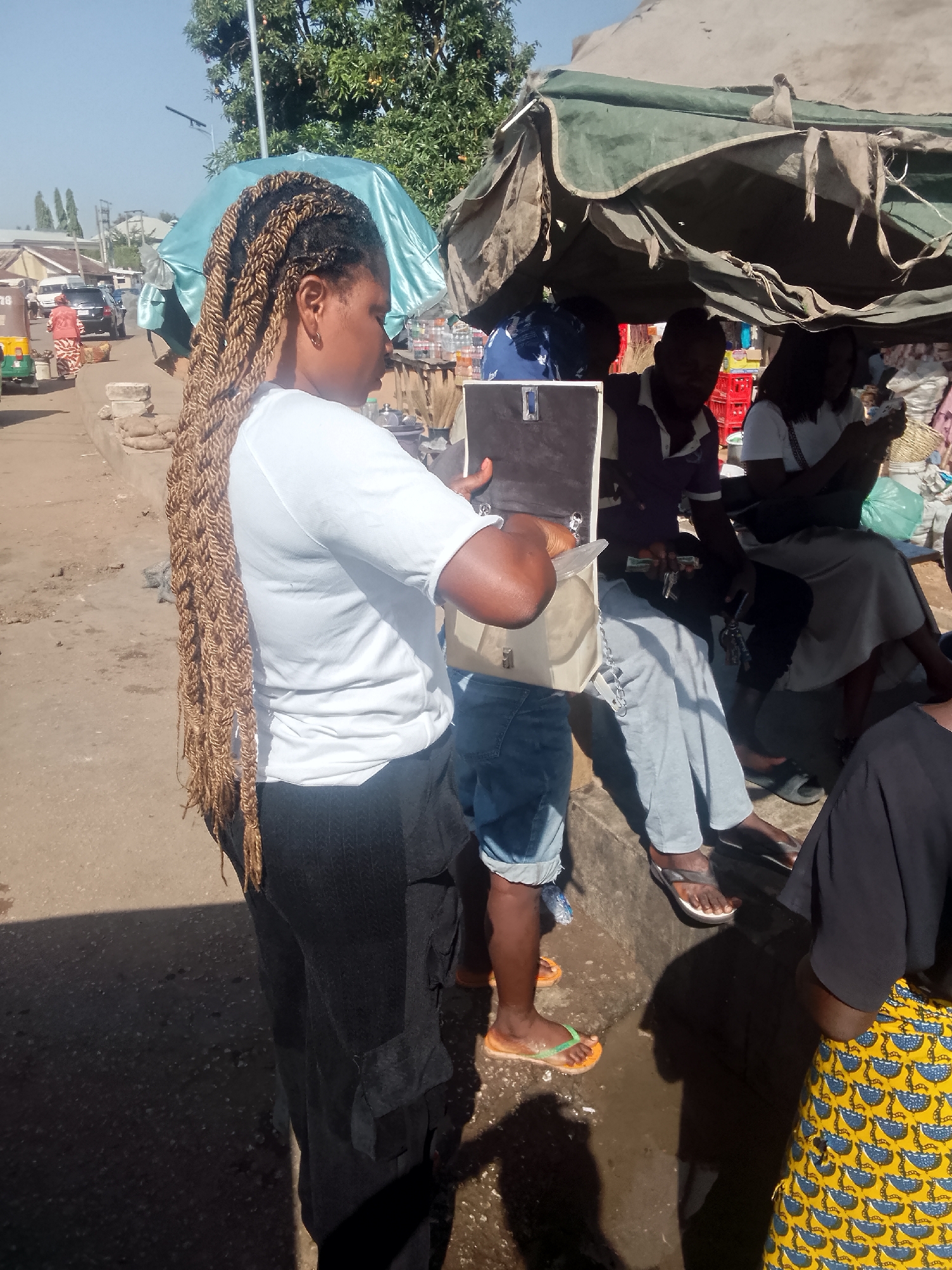 The image captures an outdoor scene in what appears to be a bustling marketplace in an unidentified city. A woman with long, intricately braided hair is the main subject, facing a group of people seated under a makeshift shelter. She is holding what appears to be a small, rectangular item or device. The people under the shelter, comprising both men and women, are engaged in conversation and activities. Overhead, an umbrella and a weathered tarp-like structure provide shade. The surrounding environment shows elements of urban activity, with vehicles and other individuals visible in the background, suggesting a dynamic and active public space.