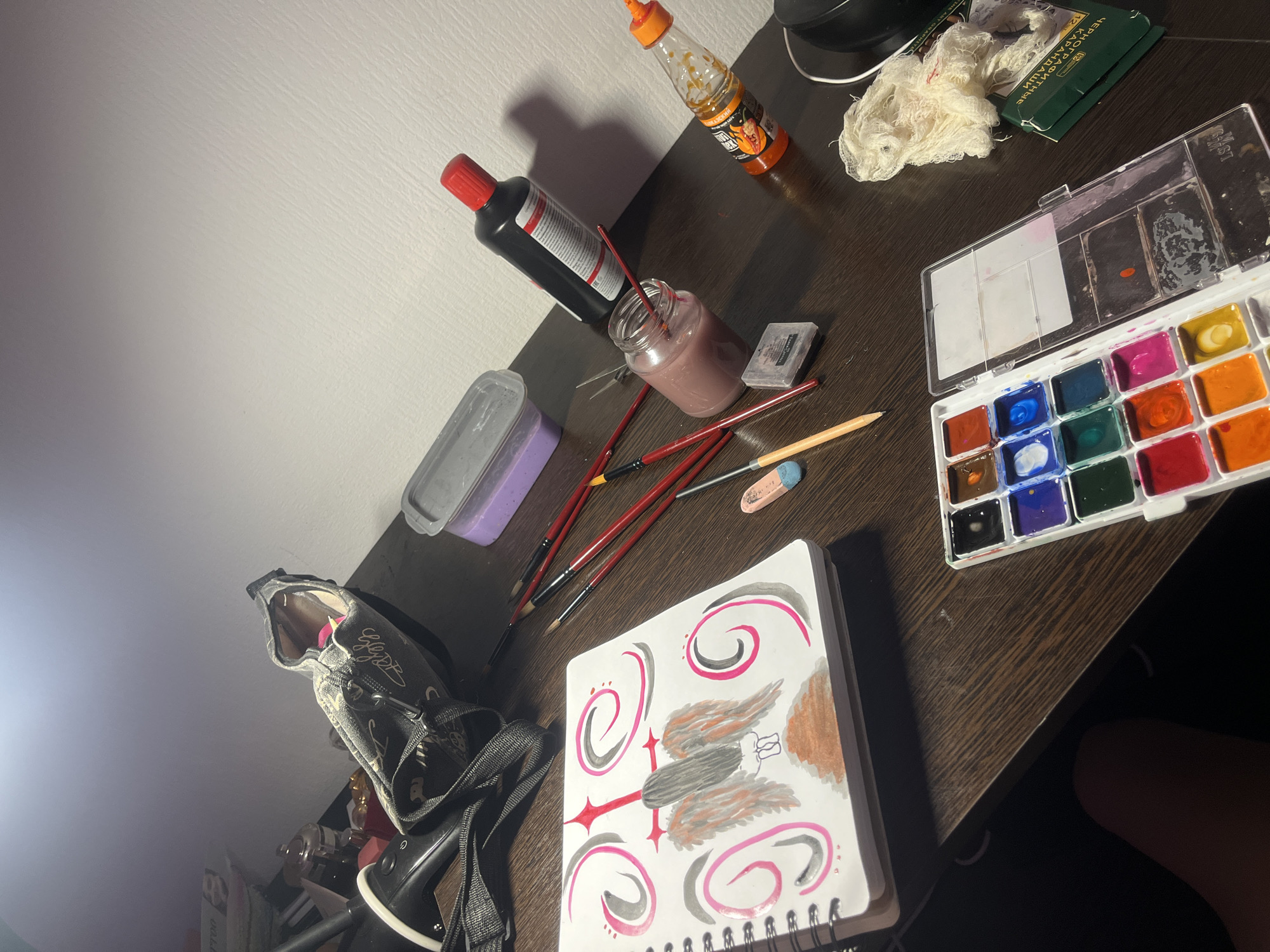 The image depicts an art desk or table with a variety of art supplies scattered across its surface. There is a sketchbook with a drawing of stylized wings and spirals in pink, gray, red, and brown. Next to the sketchbook are several paintbrushes, an eraser, and a purple container. A jar containing red paint, a bottle of black paint, and a paint palette with various colors are also present. Additional items include a bottle of hot sauce, a box, and a crumpled cloth. A shoe is visible near the left edge of the table. The table has a dark brown wood grain surface.
There is no indication of the location of the photo in Saint Petersburg, Russia.