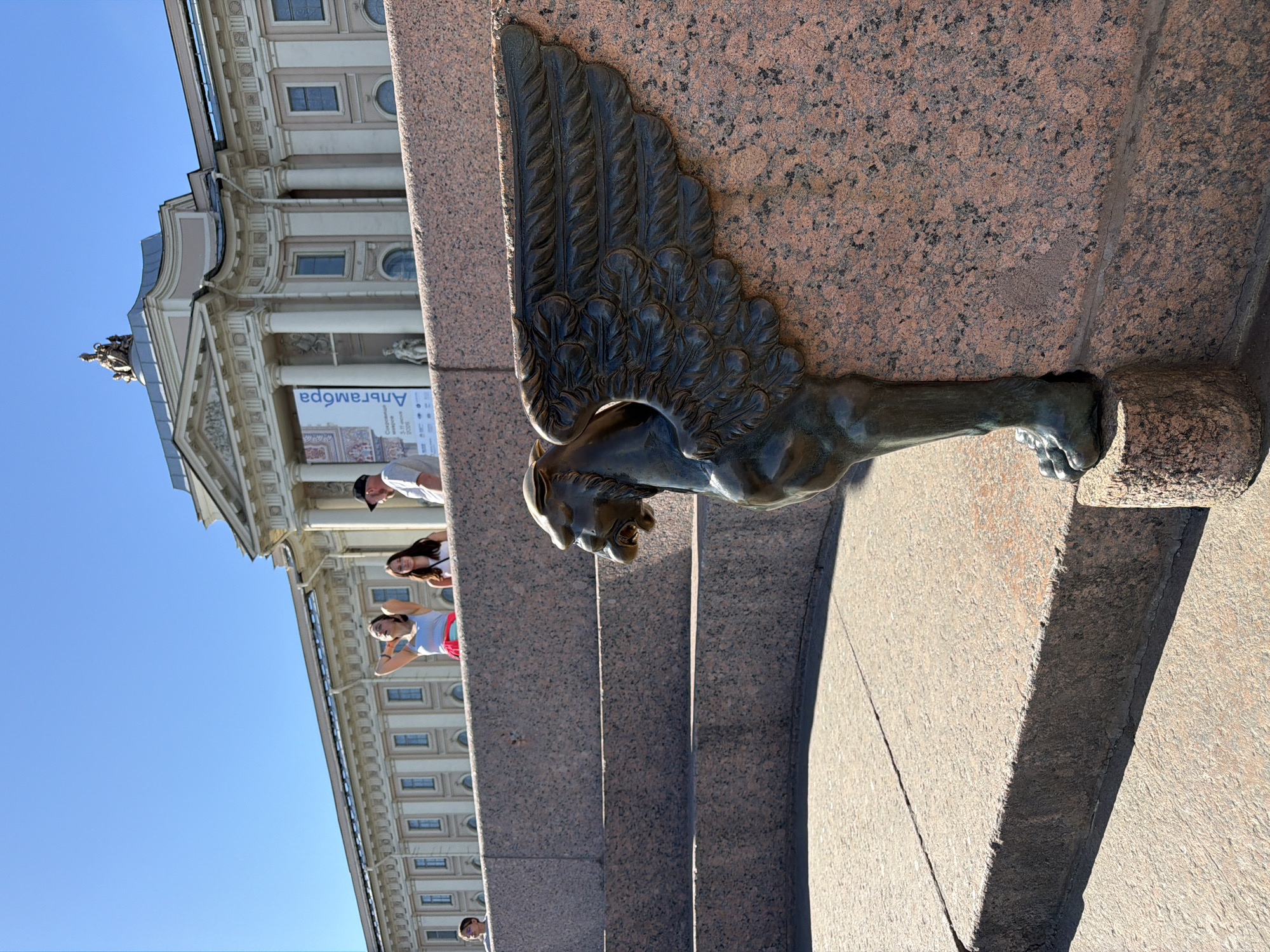 The image captures a vibrant architectural scene in Saint Petersburg, Russia, on a clear, sunny day. In the immediate foreground, a striking bronze griffin, a mythical creature with the body of a lion and the head and wings of an eagle, is prominently featured. Its dark, intricate wings are detailed with feathers, and it appears to emerge from a large, speckled reddish-brown granite structure, possibly a monument or a bridge pillar. The griffin's powerful, clawed foot is embedded in the granite.
Behind this impressive sculpture, a grand, multi-storied classical building dominates the mid-ground. It features numerous columns, arched and rectangular windows, decorative cornices, and a statue adorning its roofline. The building's light-colored facade contrasts with the deep blue sky.
Several people are visible in the vicinity of the granite structure and the building, appearing to be tourists or onlookers. One woman in a white top and red shorts is seen with her hands behind her head, looking towards the griffin. Other individuals, including a man in a white shirt and dark cap, are partially visible, suggesting a lively public space.
A blue and white banner, inverted in the original image, is displayed on the building. It shows the Cyrillic text "АЛГАМБРА" (Alhambra) and smaller text indicating "5-9 июля 2008" (5-9 July 2008), likely advertising an exhibition. The low-angle shot emphasizes the grandeur of the architecture and the detailed sculpture, set against a bright, clear sky, indicating pleasant weather, possibly summer. The griffin on a granite base strongly suggests the Anichkov Bridge, a famous landmark in Saint Petersburg.