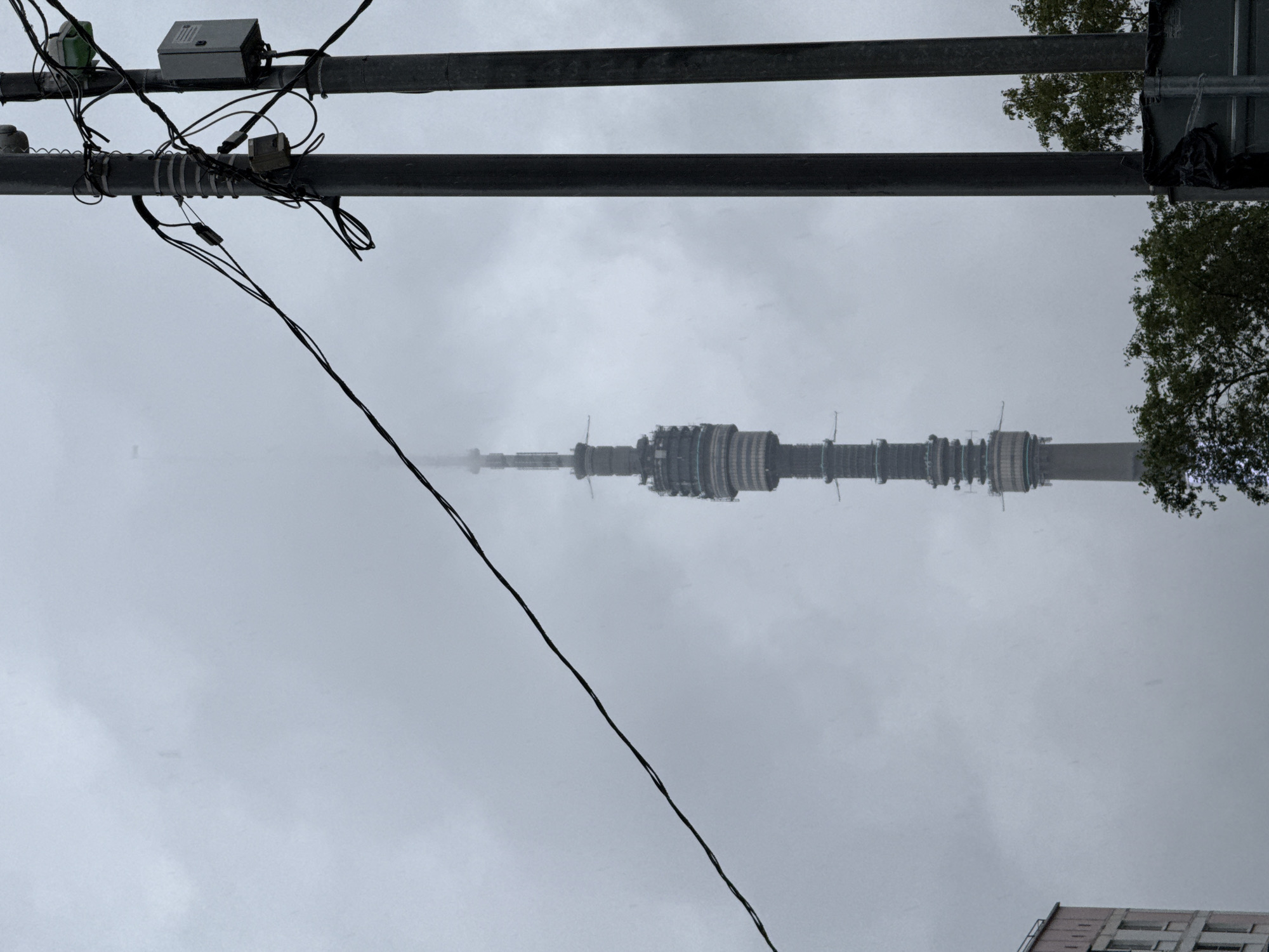 The image depicts a view of the Ostankino Tower in Moscow, Russia, on a cloudy day. The tower, a prominent landmark, is partially obscured by fog and clouds. In the foreground, two dark utility poles with numerous wires and electrical boxes are positioned, creating a visual barrier. A portion of a modern building with a pinkish facade and multiple windows is visible on the far left of the frame. Green foliage from trees is present at the bottom right, extending upwards. The overall atmosphere is subdued due to the overcast weather.