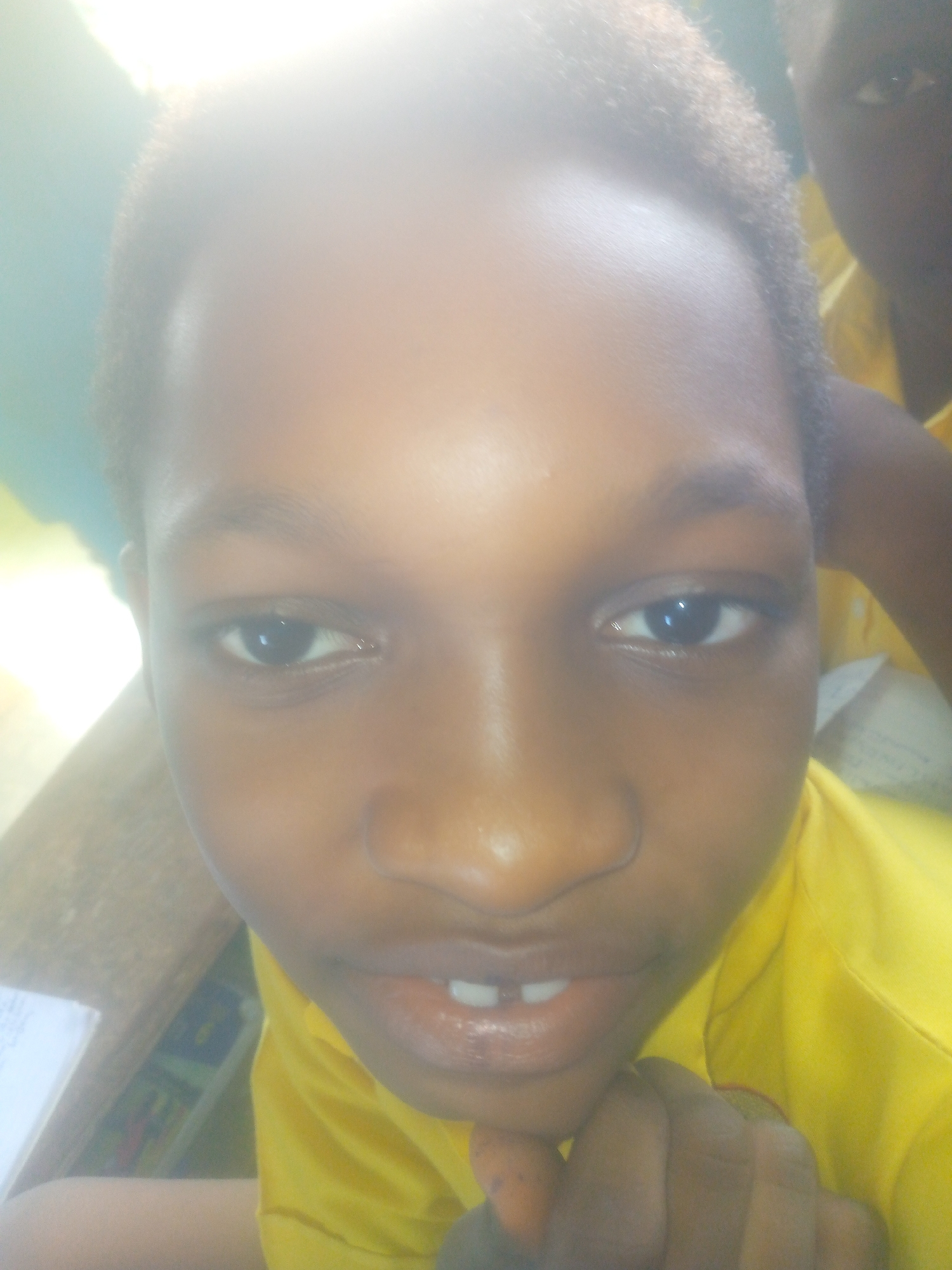A close-up selfie-style photograph shows a young person with dark skin wearing a yellow shirt. The person's face is prominently displayed, with eyes looking slightly to the side, and a partial view of teeth. The photograph appears to be taken indoors. A portion of another person's face and yellow shirt can be seen in the upper right corner of the image. The photo does not contain any information to identify the city of Ikorodu or Nigeria. The location is based on provided information and not visible within the image.