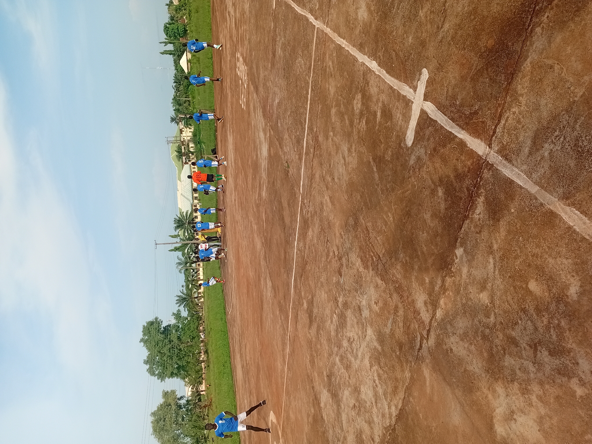 The image depicts a group of individuals on a brown, dirt field with white lines, likely a sports field, in Ogbogbo, Nigeria. A line of individuals wearing blue shirts with visible numbers are lined up along the field, possibly in preparation for a game or drill. One individual wears an orange shirt. Some of the individuals are in motion, suggesting active participation. The environment includes green grass, trees, a building, and a blue sky. A white "X" is visible on the ground.