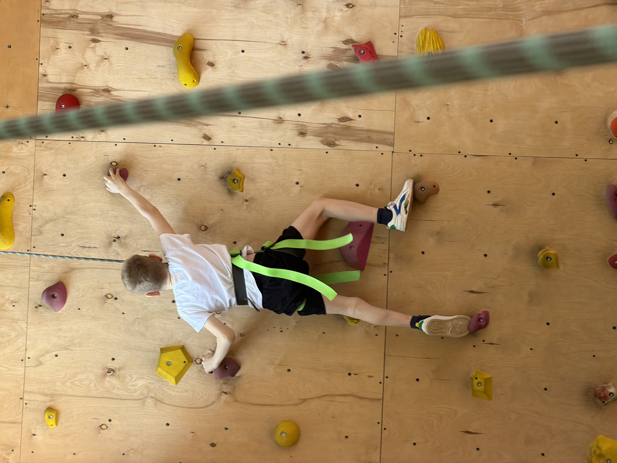 A young boy is captured in a top-down, slightly angled view while actively rock climbing on an indoor climbing wall in Izhevsk, Russia. The boy, with short brown hair, is wearing a white short-sleeved t-shirt, black shorts, and a bright light-green climbing harness. His body is positioned horizontally across the light-colored wooden wall, which is constructed from multiple panels and features numerous small screw holes and seams.
The wall is adorned with a variety of brightly colored climbing holds in different shapes, including solid yellow, red, purple, orange, and distinctive hexagonal yellow holds. The boy's left hand is gripping a red hold, and his right foot is firmly planted on a purple hold, indicating active movement during his climb. His limbs are extended, showcasing his engagement with the sport. A thick, green and light-green striped safety rope is prominently featured, running diagonally across the image in the foreground, appearing slightly out of focus, and is connected to the boy's harness. He is wearing athletic shoes, with the right shoe displaying blue and yellow accents. The setting is an indoor climbing facility, identifiable by the specialized equipment and consistent lighting. No text is visible in the image, and the boy's focused posture suggests concentration on his activity.