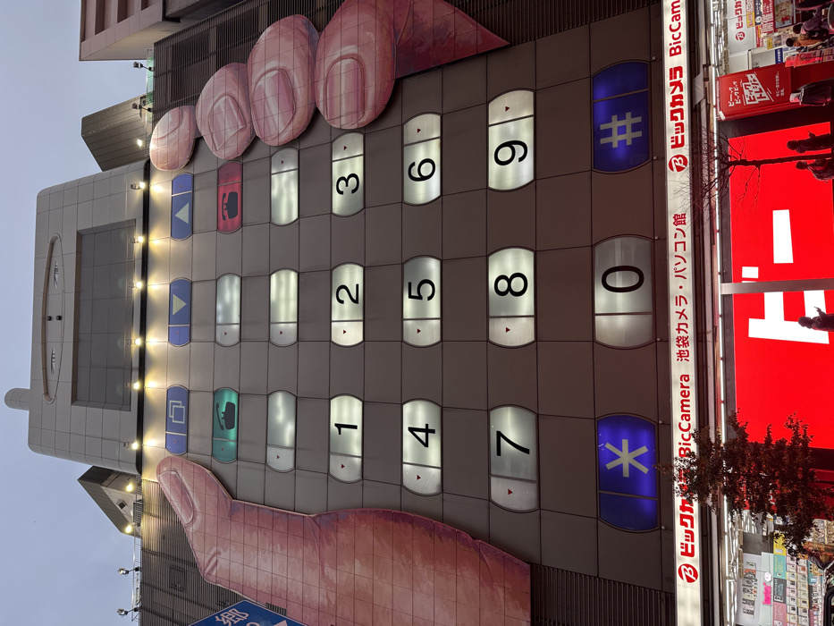 This image shows the facade of a building designed to resemble an oversized mobile phone. A giant hand graphic is present, with fingers depicted as pressing the numerical keypad. The buttons are arranged in a typical phone layout, with numbers 1 to 0, plus asterisk and hash symbols. Adjacent to the building is signage for Bic Camera, indicating the store's location in Ikebukuro, and advertising its camera and computer products.