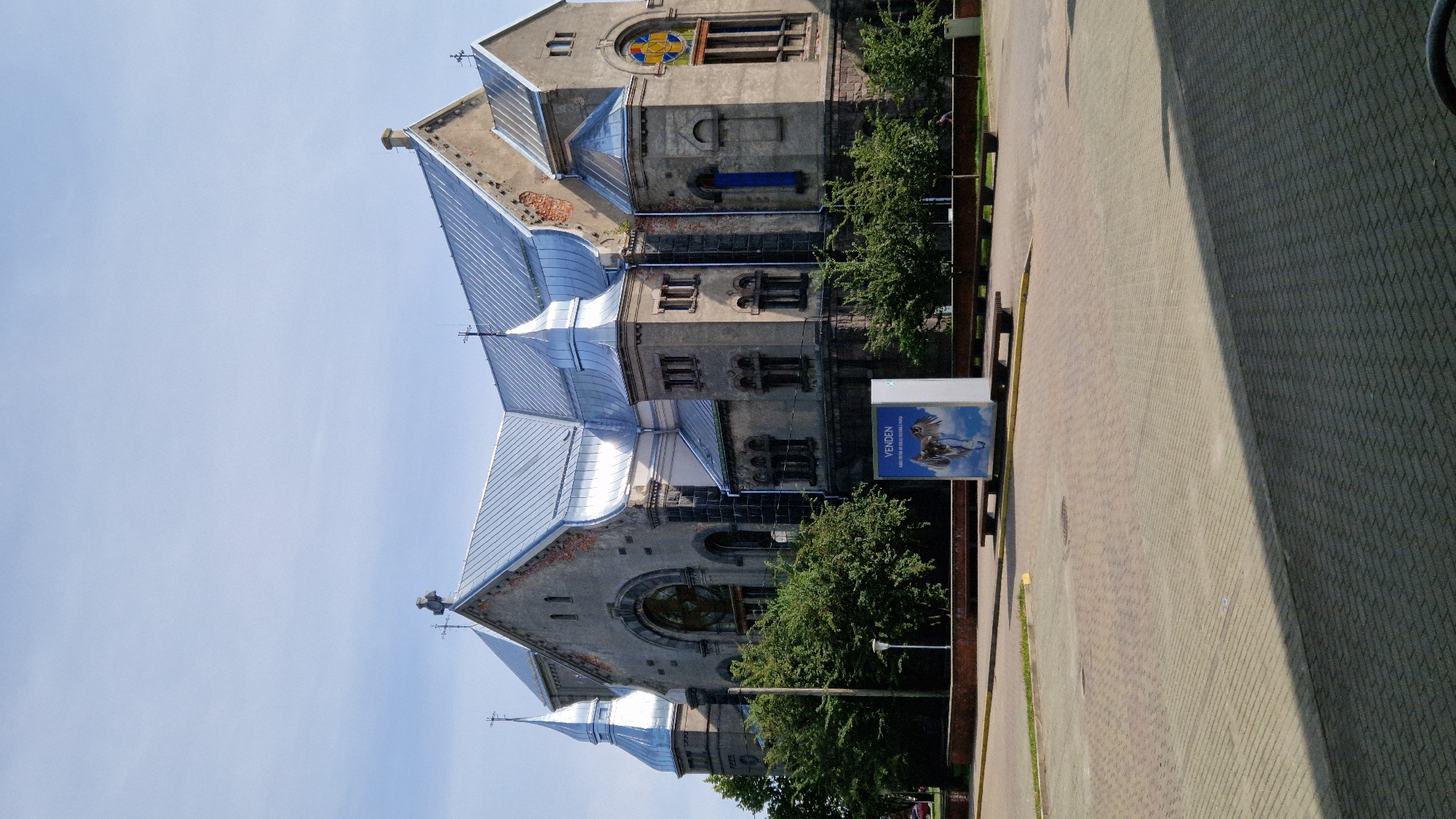 The image captures a sunny daytime view of the Liepaja Evangelical Lutheran Great Charity Church in Liepaja, Latvia. The church, constructed of stone and featuring a prominent silver metal roof, is the central subject. It exhibits architectural details such as multiple towers with spires topped by crosses, arched windows, and decorative stonework. A large circular window with stained glass is visible on the right side of the building, and a rectangular stained-glass window is present on the far right. Lush green trees surround the church. In the foreground, a paved plaza with a brick-like pattern is visible, leading to a low wall and benches. A modern advertising stand with an image of two winged horses and text is placed in front of the church, to the left of the center. A single person is visible in the distance on the right side of the plaza, walking. The sky is clear blue with a few faint clouds.
